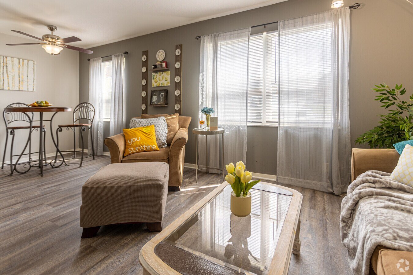 Bright living room with large windows, sheer curtains, a beige sofa with yellow and white pillows, a glass-topped coffee table with a flower vase, a small side table with a flower arrangement, a wall decoration with plates, and a dining area with a table and chairs.