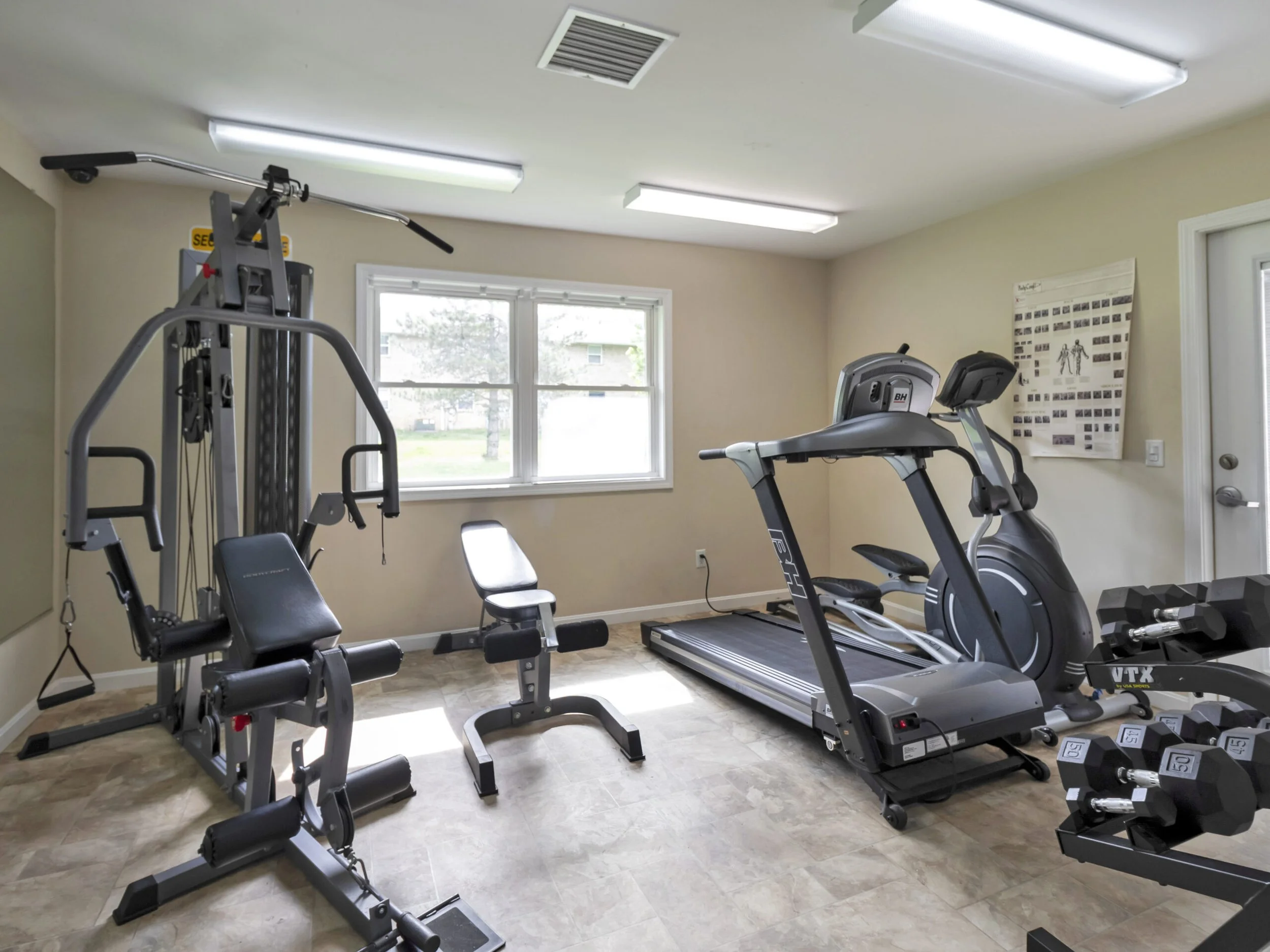 A small home gym with a treadmill, elliptical machine, weight machine, and free weights. There are large windows with natural light and a poster on the wall.