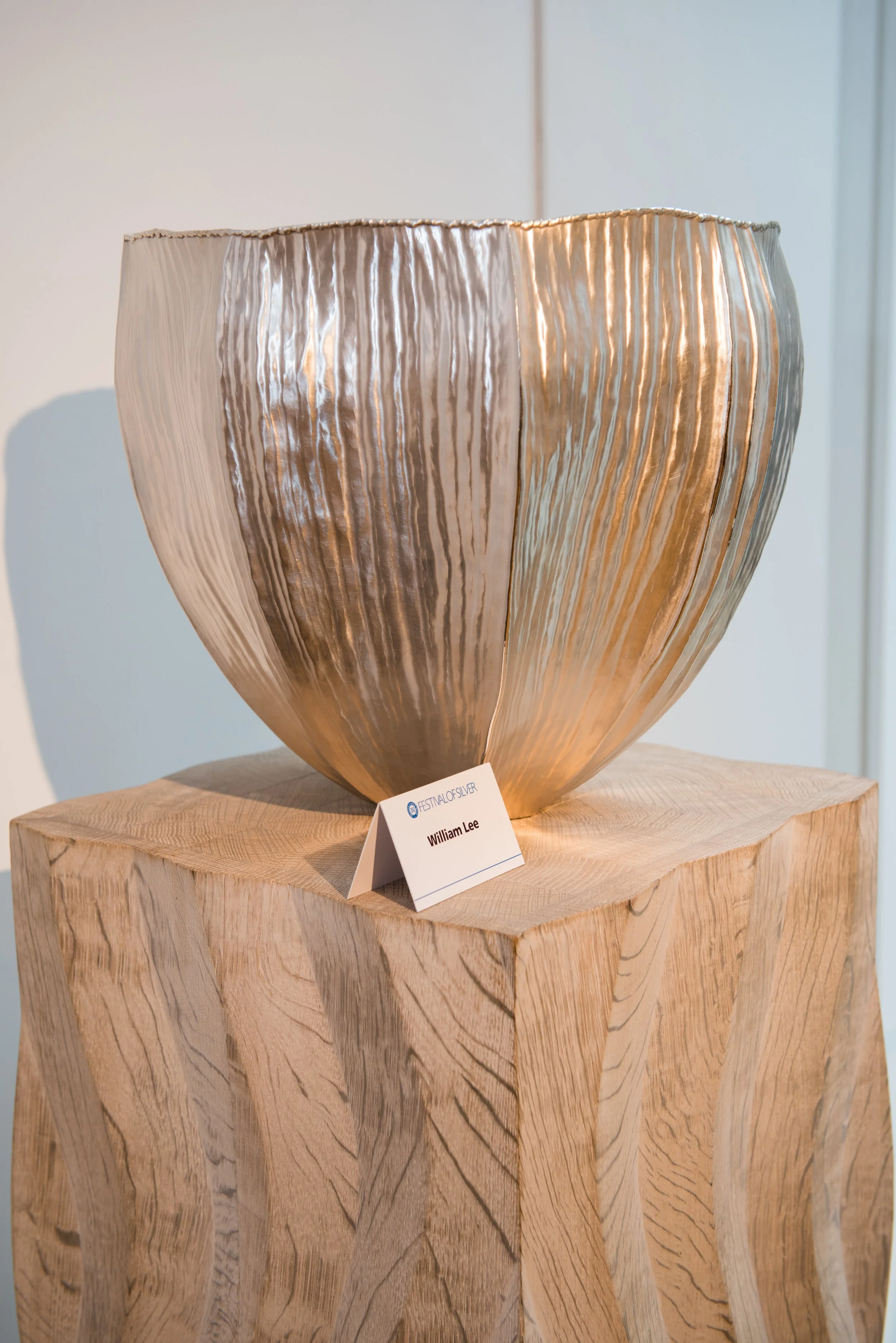 A large, metallic, flower-shaped sculpture with wavy engraved lines, displayed on a wooden pedestal at an art exhibition.