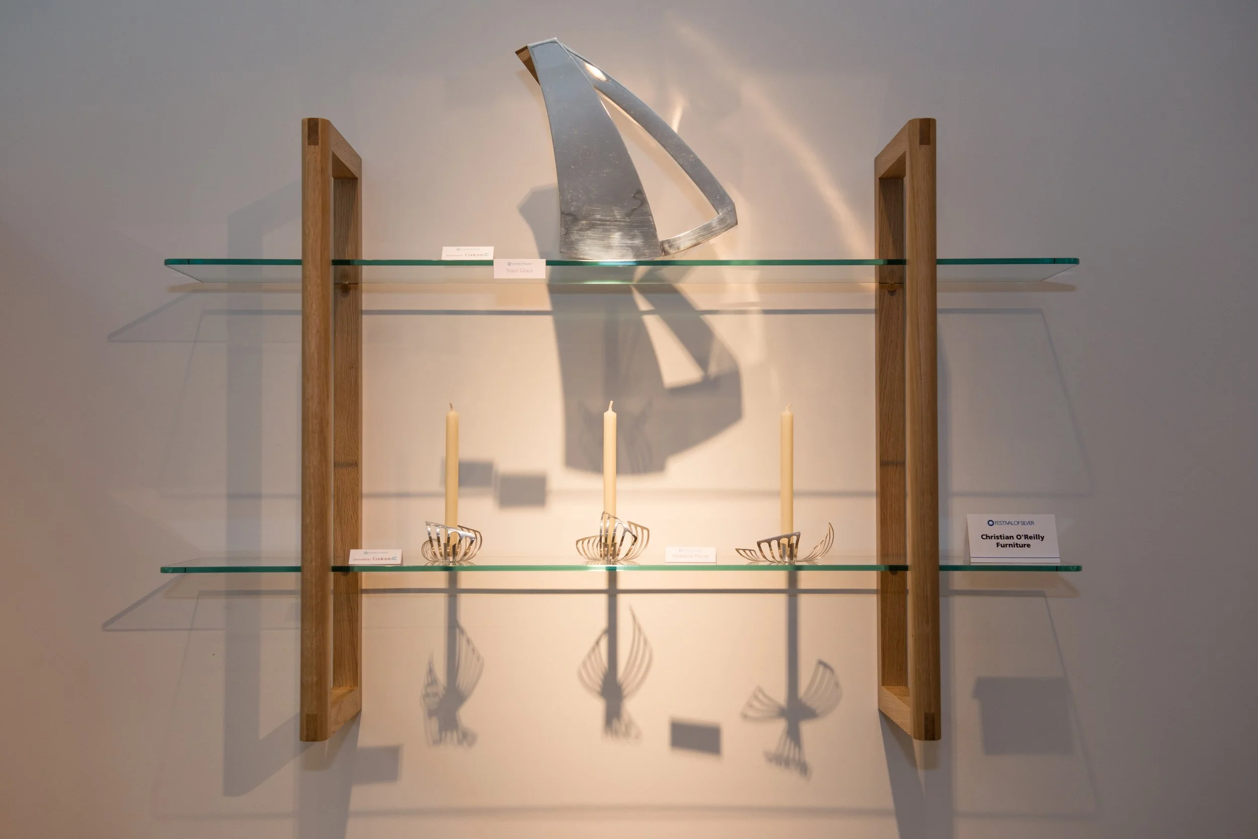 Display shelf holding modern silver furniture pieces with intricate wire designs, wood supports, and candles, in a gallery setting.