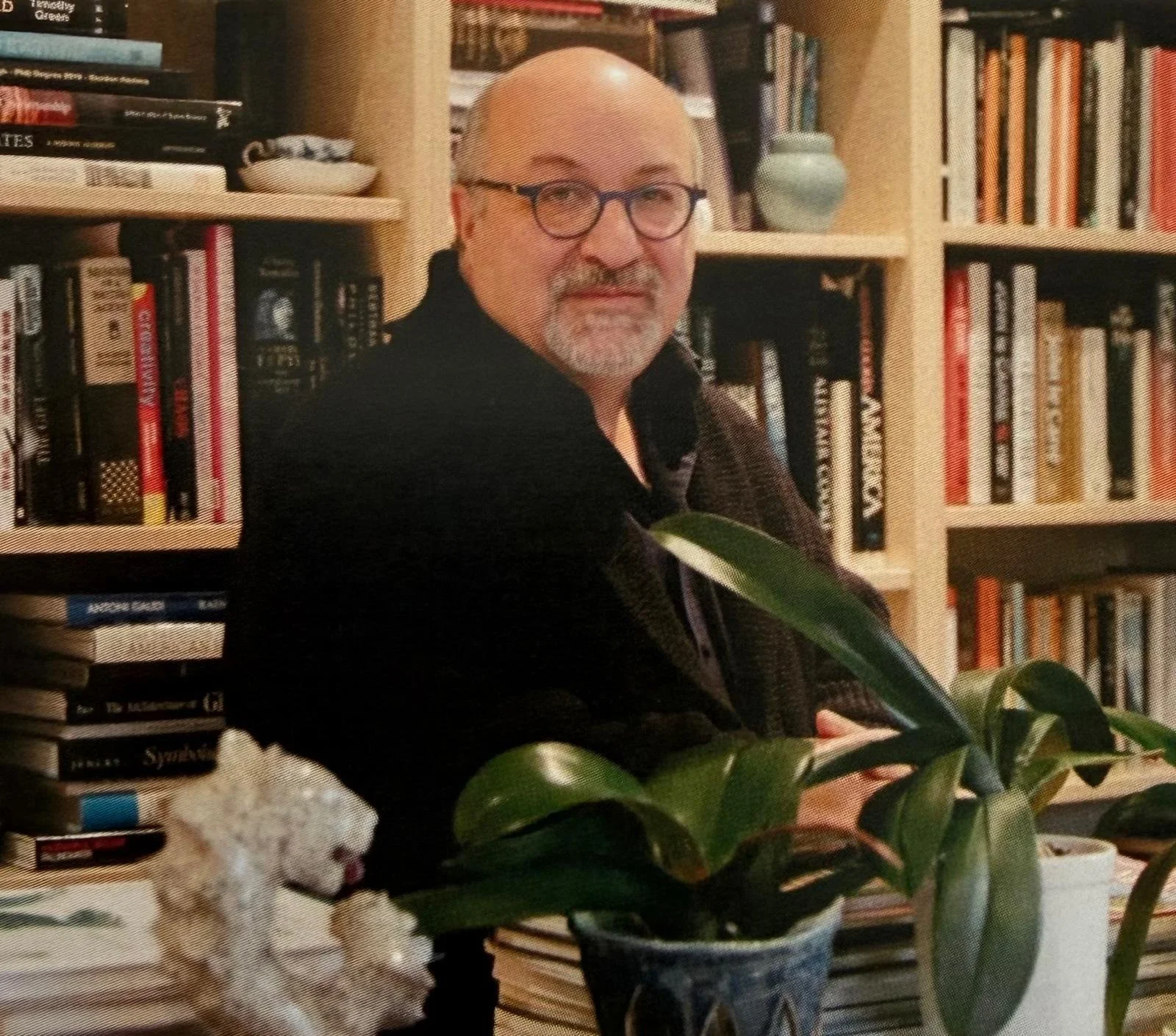 An older man with glasses and a white beard, smiling, is sitting in a room with wooden bookshelves filled with various books in the background. There are large green houseplants in front of him and to the side. A small fluffy dog with white and tan fur is sitting on a table in the foreground.
