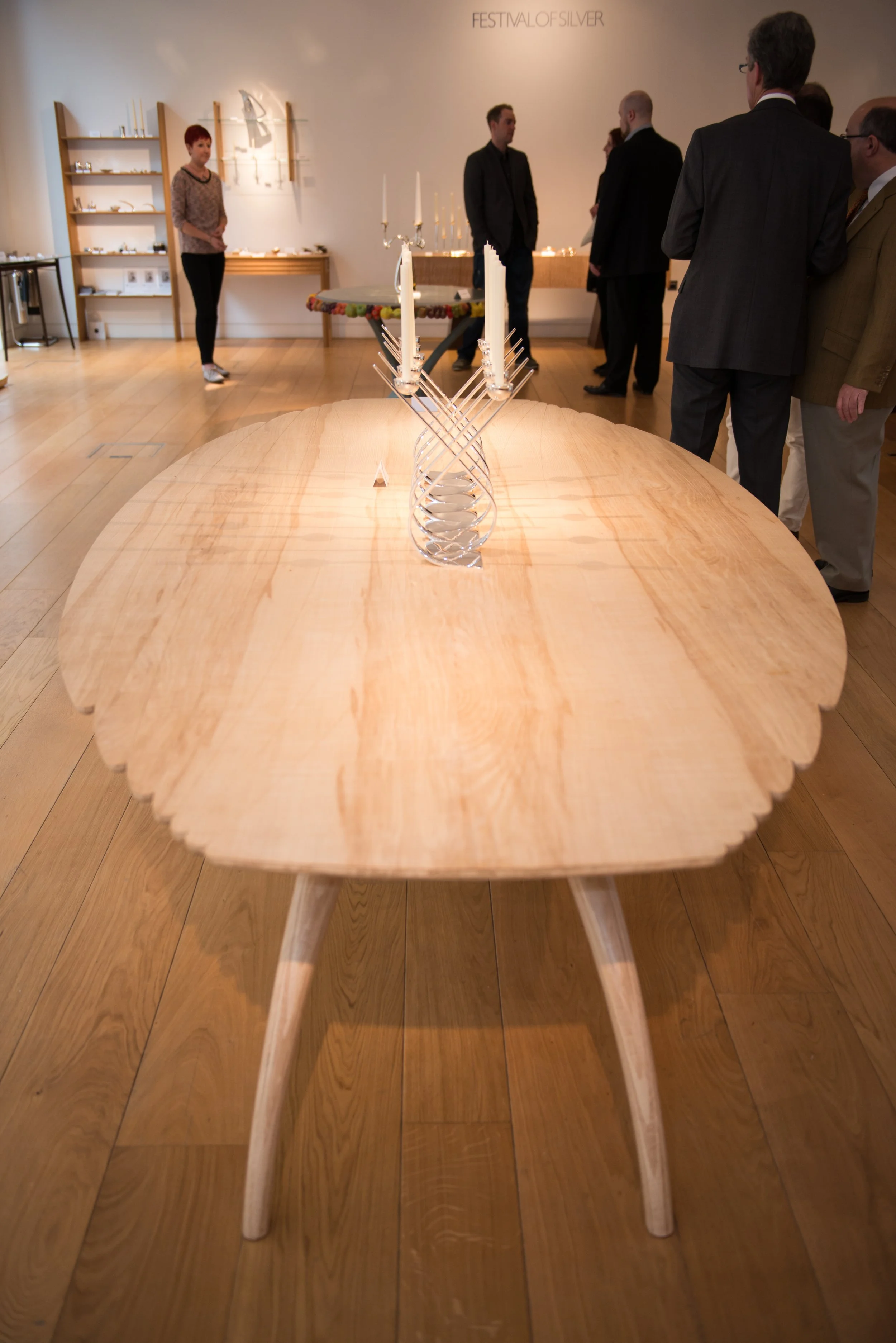 A wooden table with a candelabra holding six candles in the center of a room during an art exhibition. Several people are gathered around, some engaged in conversation, with a display shelf and artwork on the wall in the background.