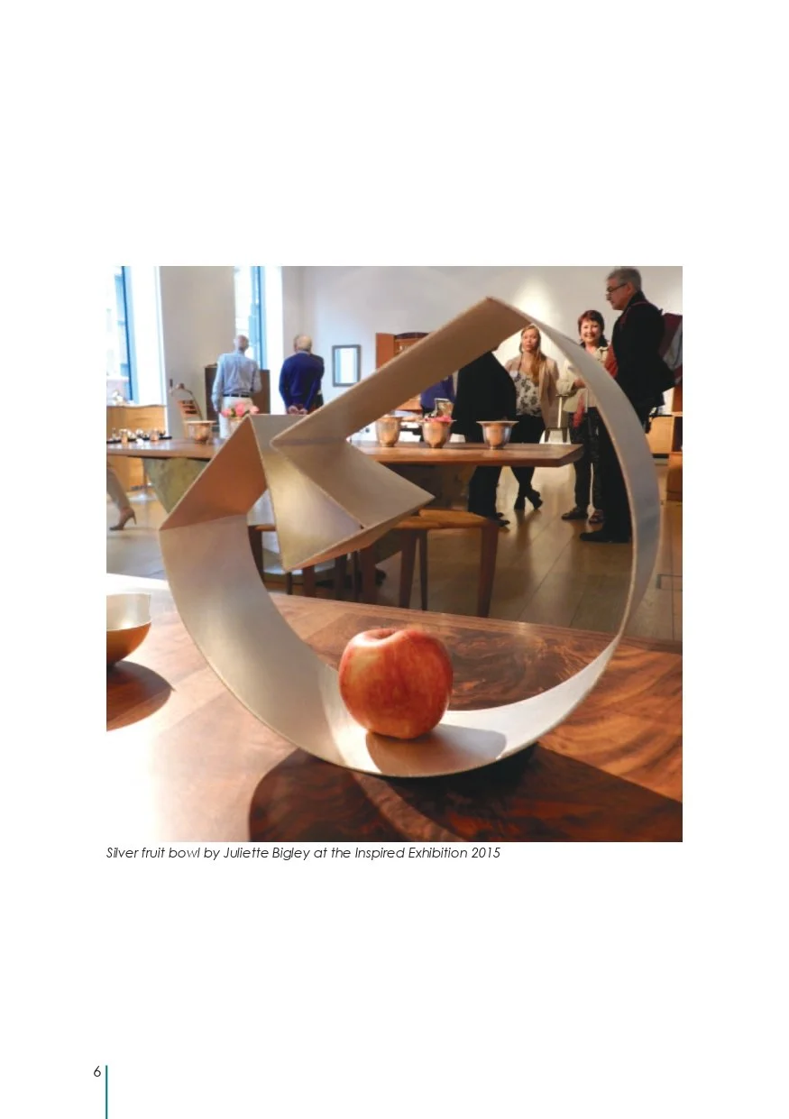 A silver fruit bowl by Juliette Bigley, with an apple inside, displayed at the Inspired Exhibition 2015, with people browsing in the background.