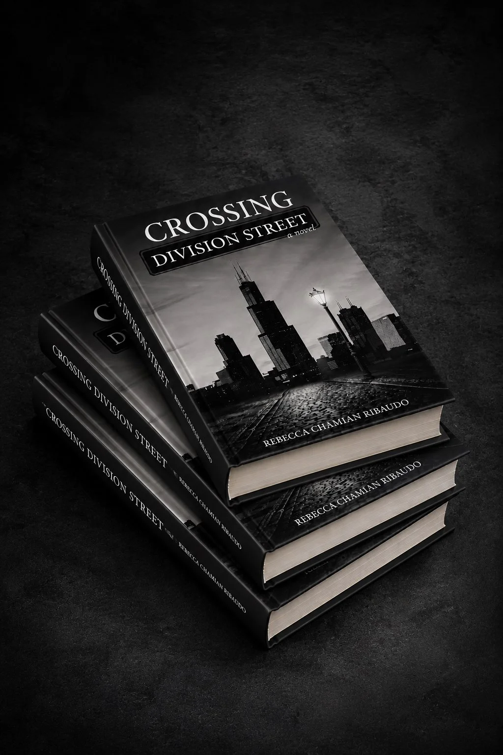Four copies of a book titled "Crossing Division Street" by Rebecca Chamian Ribaudo stacked on a dark surface, with a black-and-white cover image of a city skyline including the Willis Tower and a streetlamp.