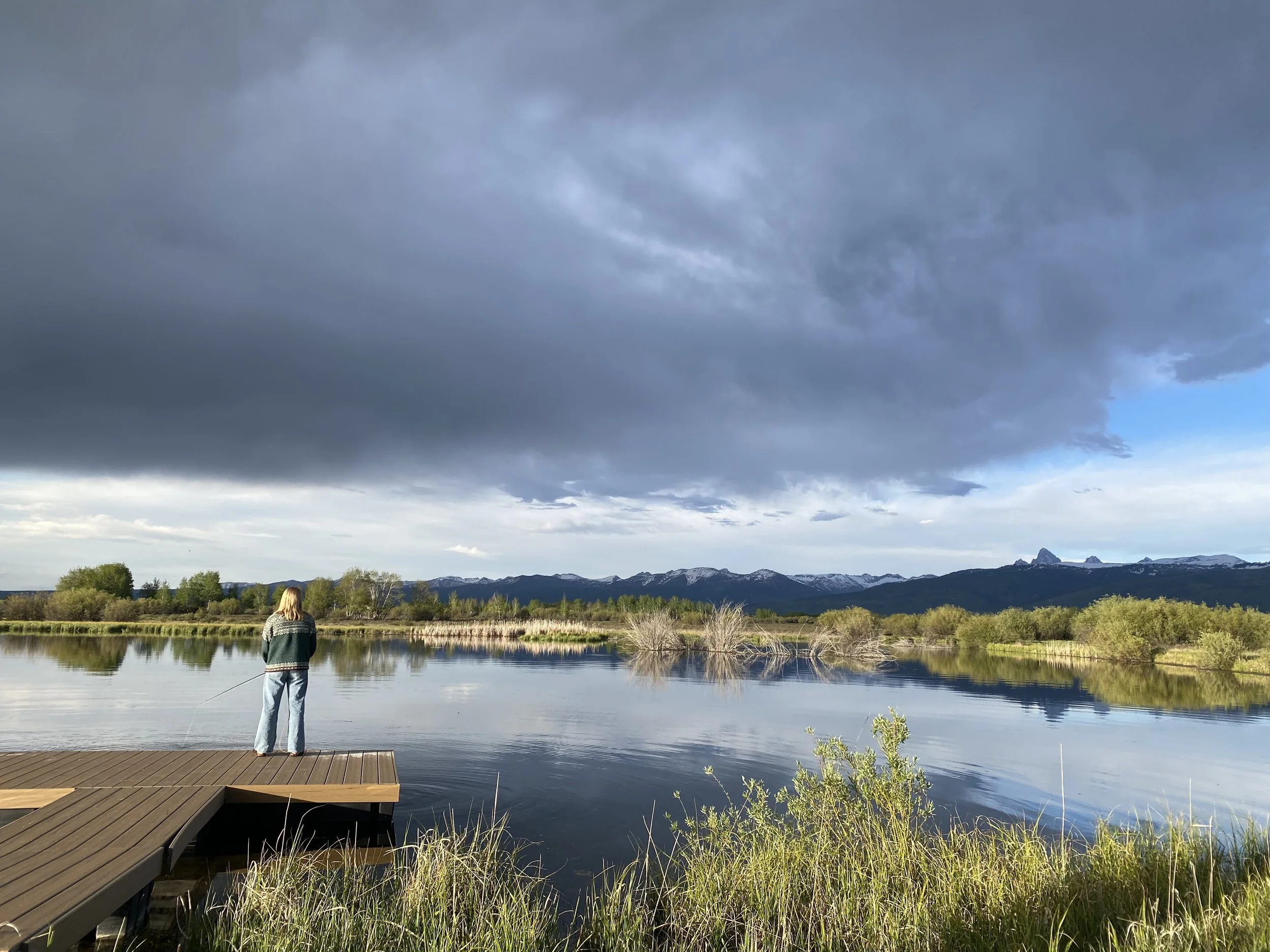 A person stands on a dock fishing by a calm river with mountains and cloudy sky in the background.