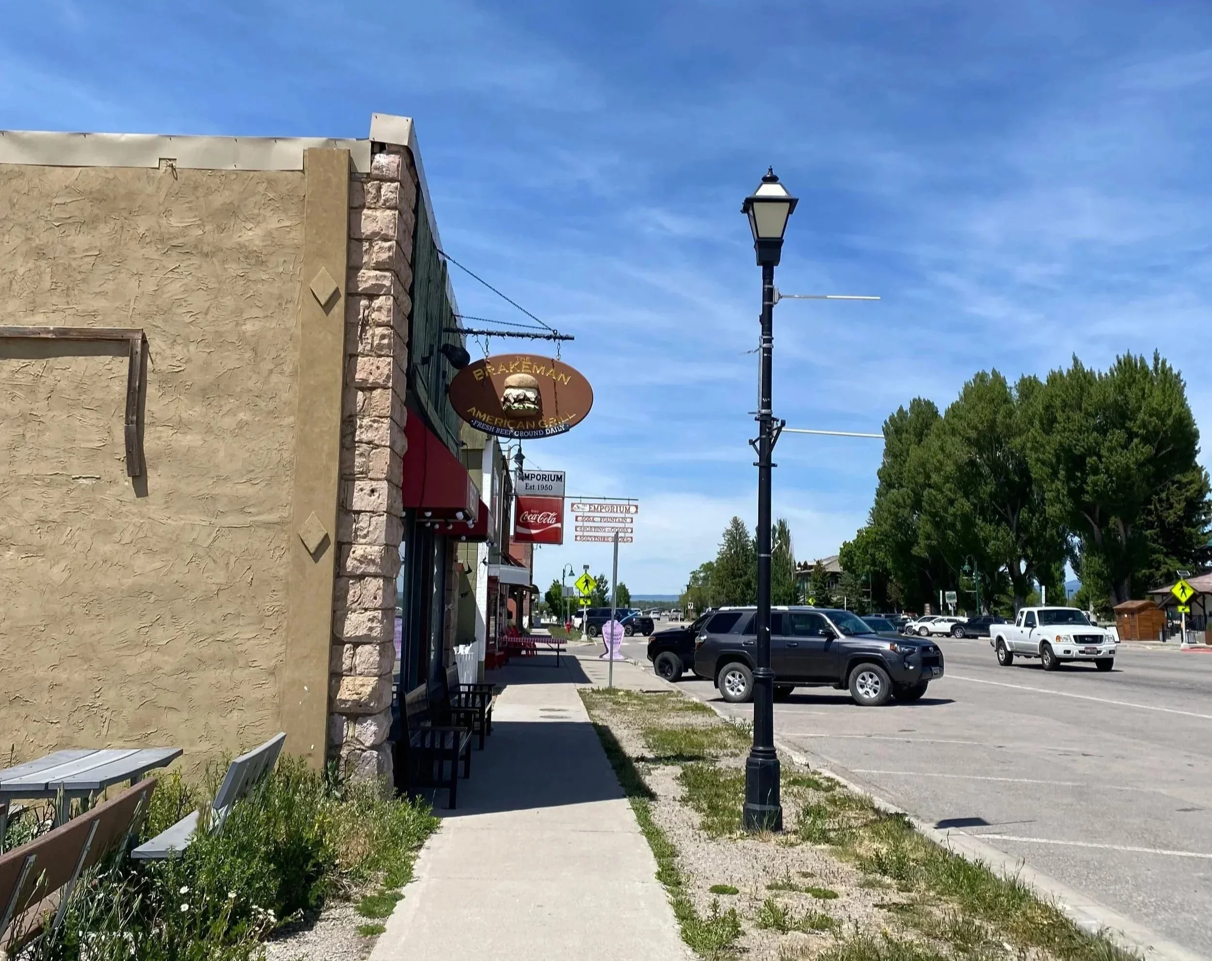Sidewalk outside a building with a decorative sign for a grill restaurant, parked cars in parking lot, street with trees, street lamp, blue sky with some clouds.
