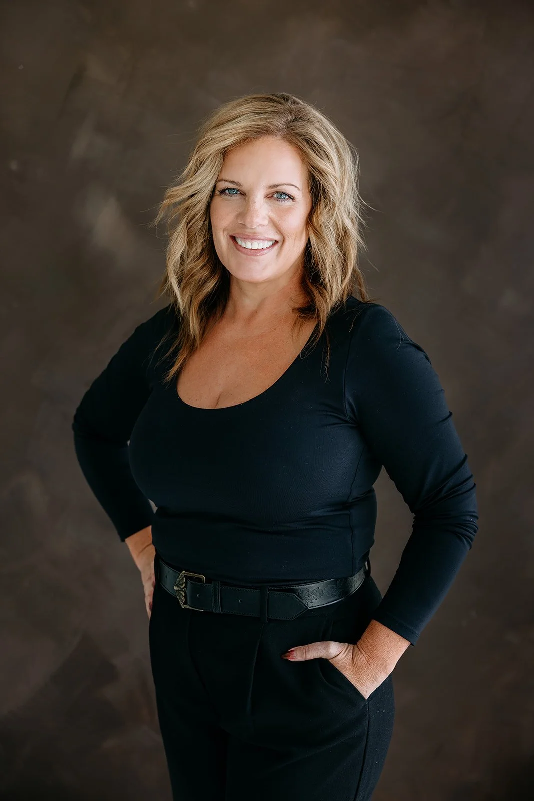 A smiling woman with wavy blonde hair wearing a black long-sleeve top and black pants, standing against a brown textured background.