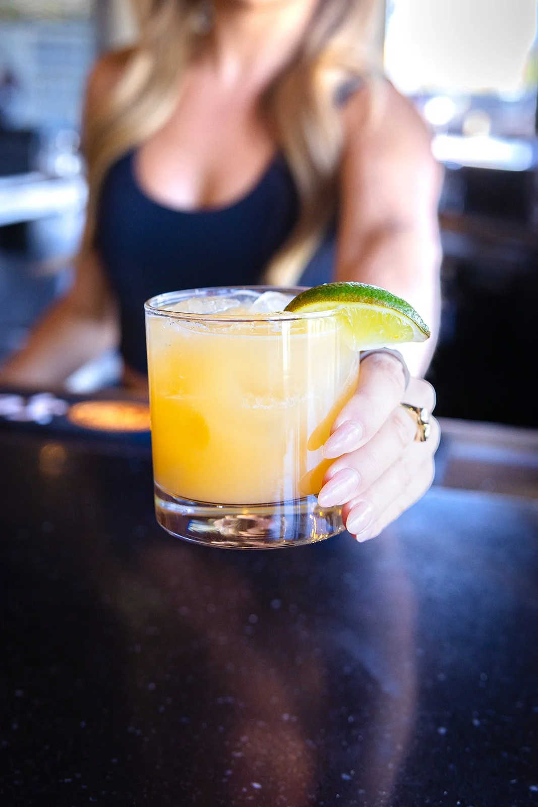 A woman holding a cocktail with a lime wedge garnishing the glass, sitting at a bar or restaurant.