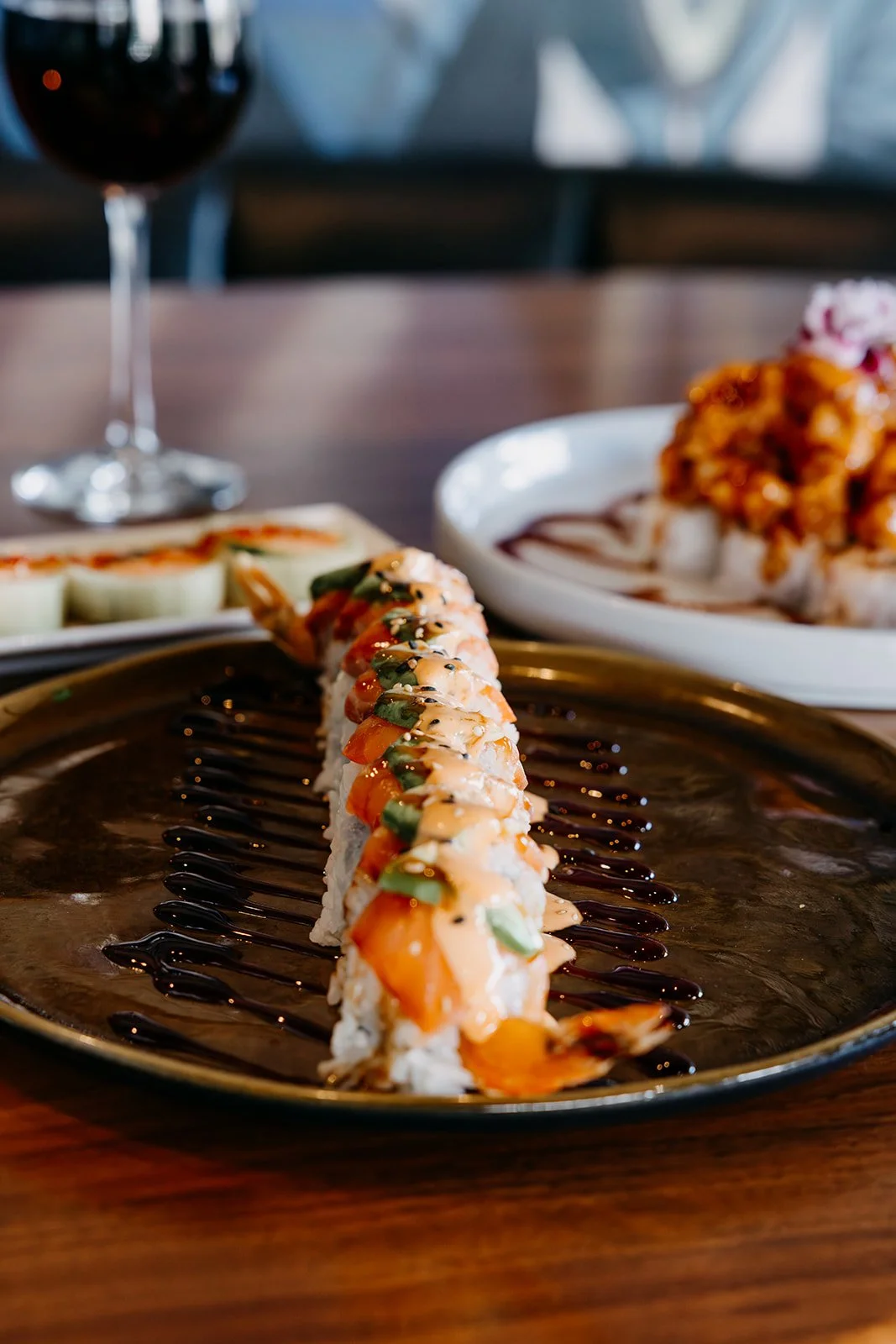 Close-up of a sushi roll drizzled with sauces on a black plate, with other sushi dishes and a glass of red wine in the background.