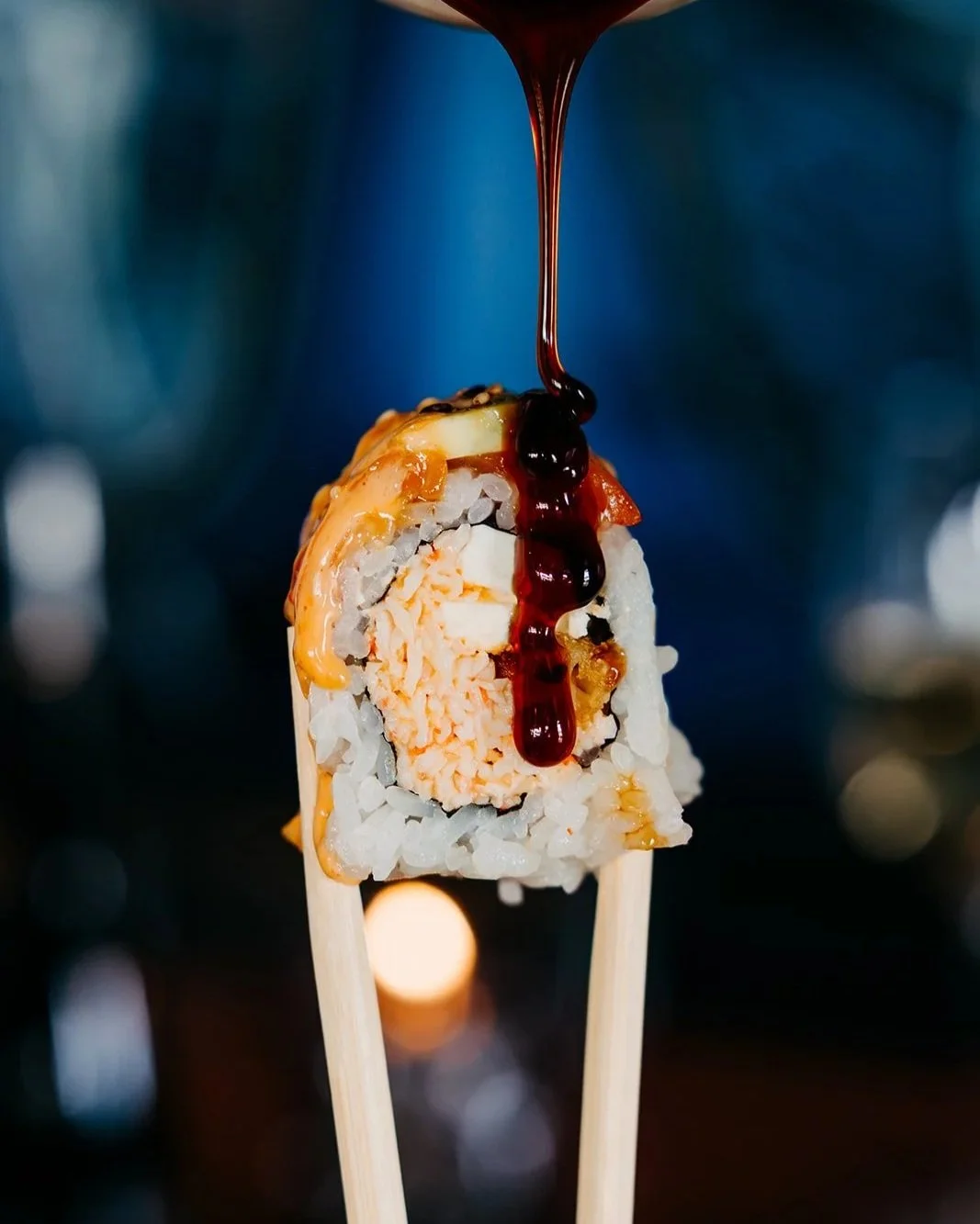 Close-up of a sushi roll with rice, fish, and cream cheese, topped with sauce and drizzled with dark sauce, held on two chopsticks with a blurred dark background.