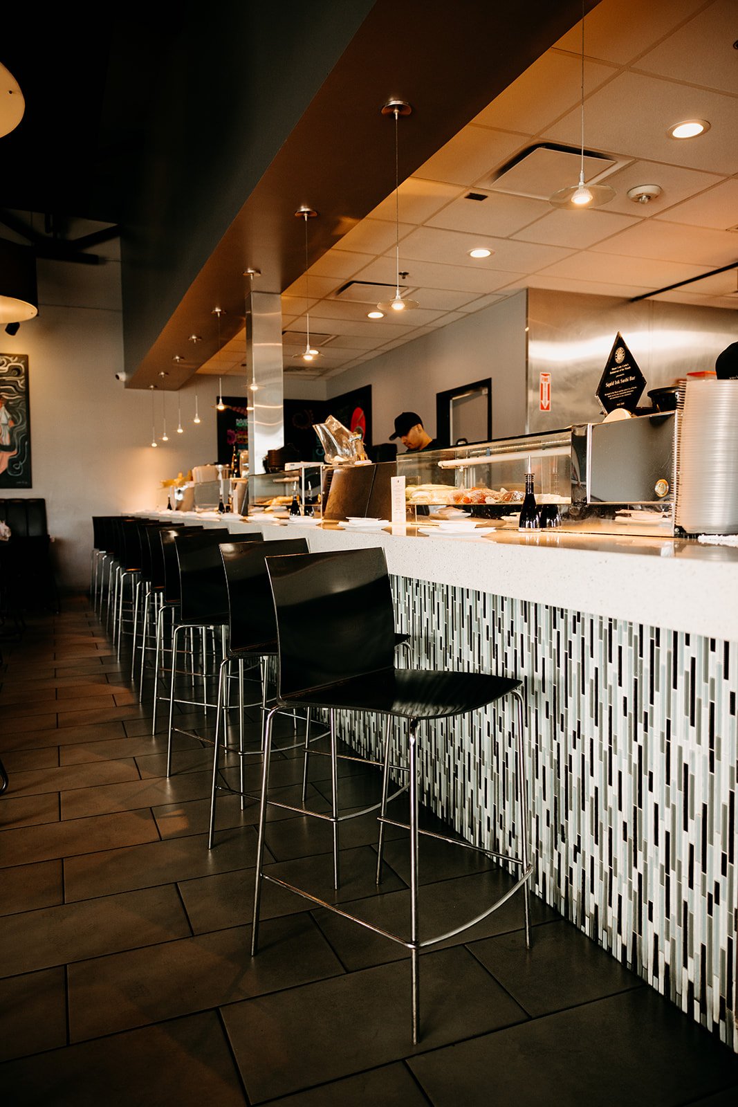 An empty restaurant with a row of black chairs at the counter, white tiled bar, and a chef working behind it, with ambient lighting and modern decor.