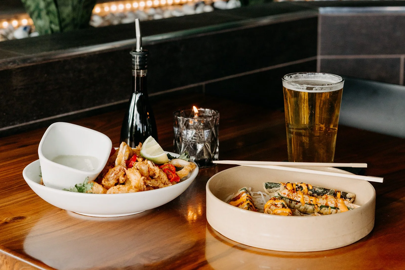 A wooden table with two bowls of Asian-style food, a glass of beer, chopsticks, a soy sauce bottle, a lit candle, and a small bowl of dipping sauce in a restaurant setting.