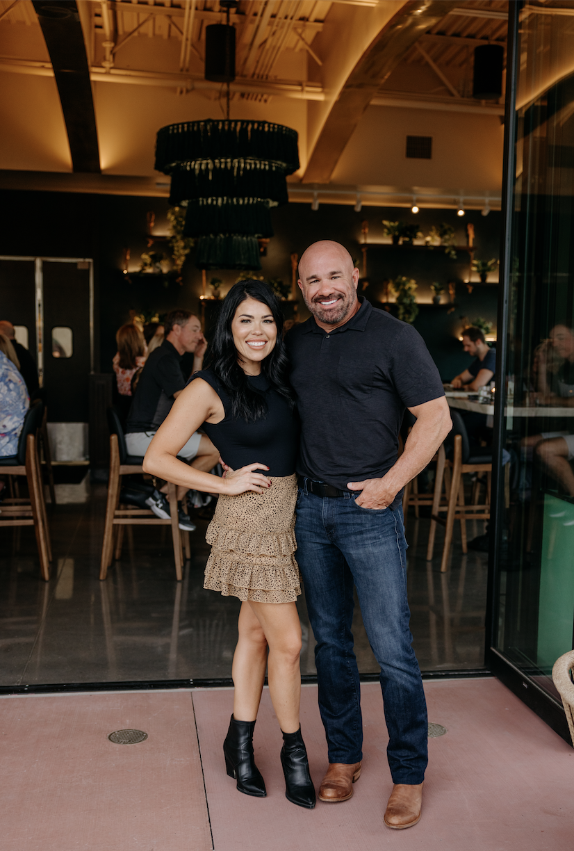 A young couple standing together, smiling inside a restaurant with people dining in the background.