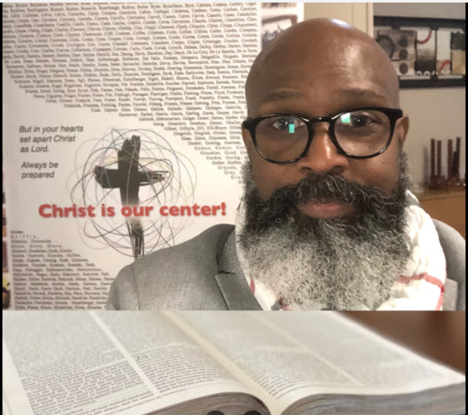 A man with glasses and a beard in a suit taking a selfie indoors, with a church or religious poster in the background that reads 'Christ is our center!'