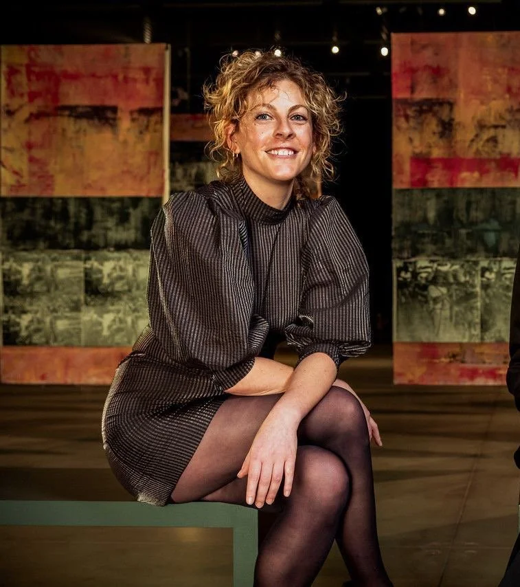 A woman with curly blonde hair, wearing a black and gray striped dress and black stockings, sitting on a bench in front of colorful abstract artwork, smiling at the camera.