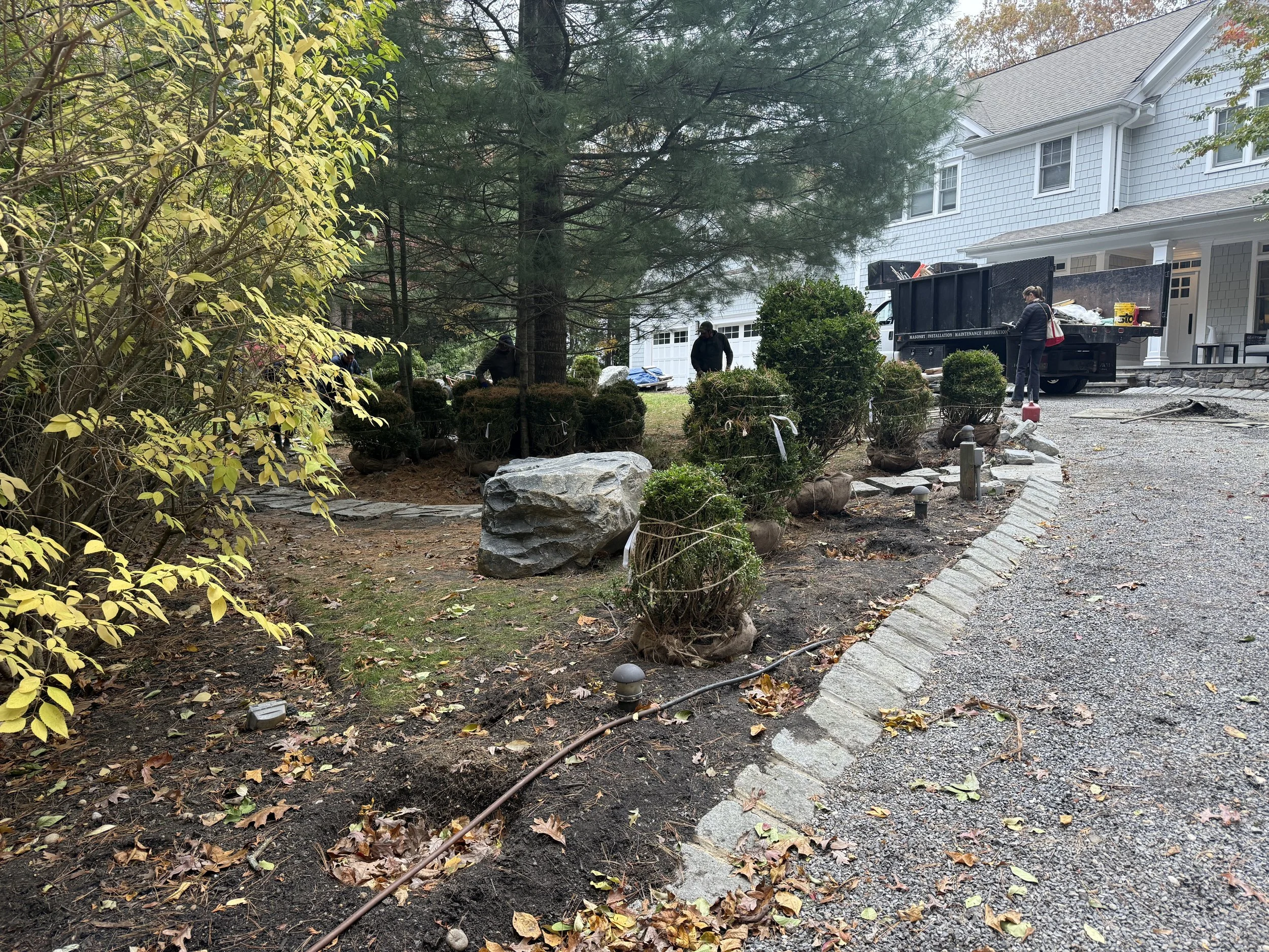 Workers landscaping a front yard with bushes, rocks, and soil near a white house with a gray roof.