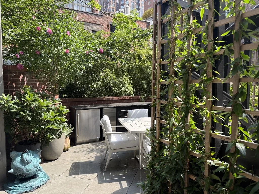 Small outdoor patio with potted plants, a decorative fish sculpture, a white table with chairs, a built-in outdoor kitchen or storage unit, surrounded by green foliage and a wooden trellis covered in climbing plants, with tall buildings visible in the background.