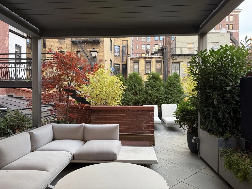 Urban apartment balcony with white sectional sofa, potted green plants, red and yellow trees, outdoor chairs, and city buildings in the background.
