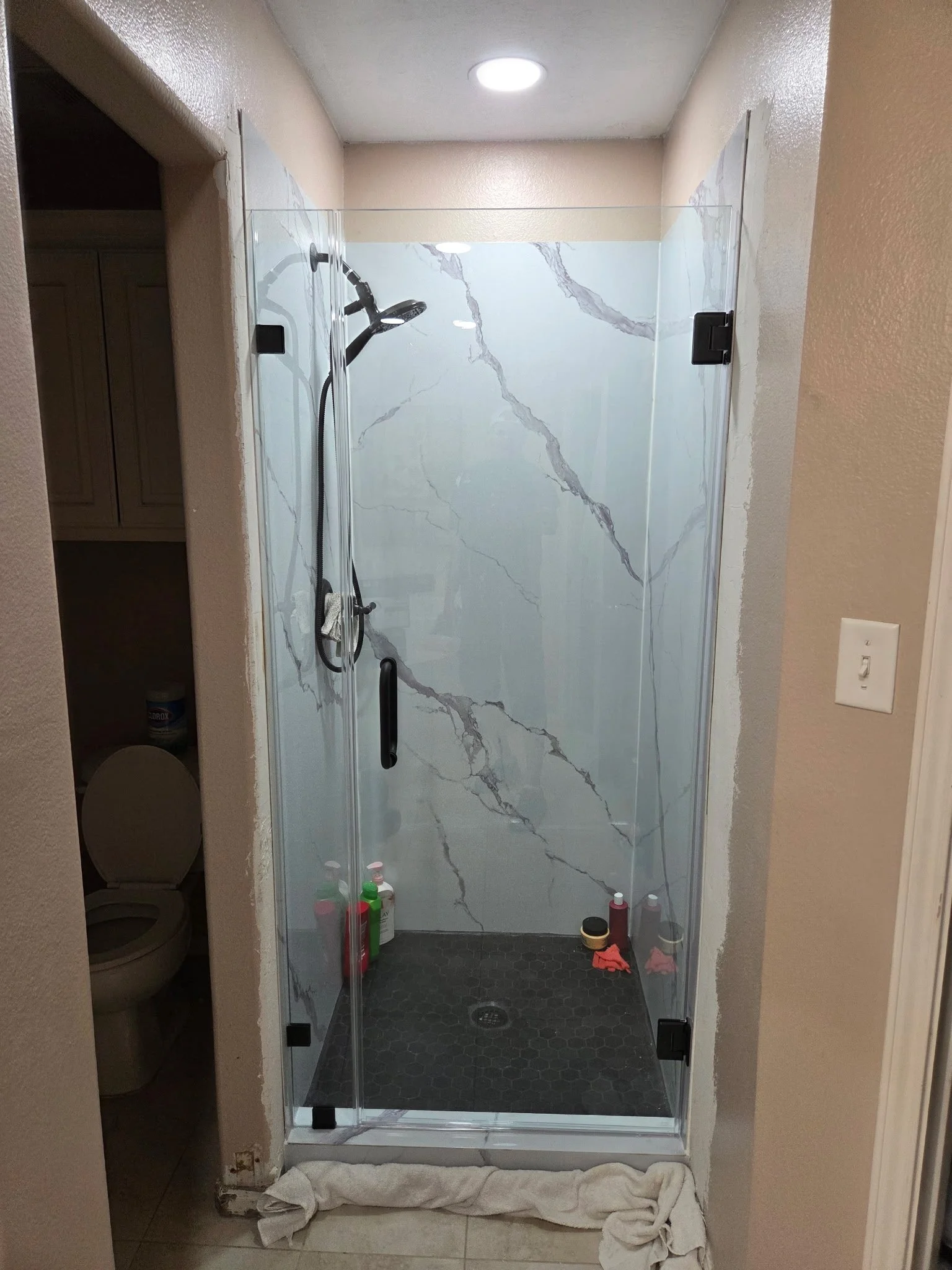 A bathroom shower with marble walls, a glass door, and various shower cleaning supplies on the floor, with a white towel or cloth spread out on the floor outside the shower.