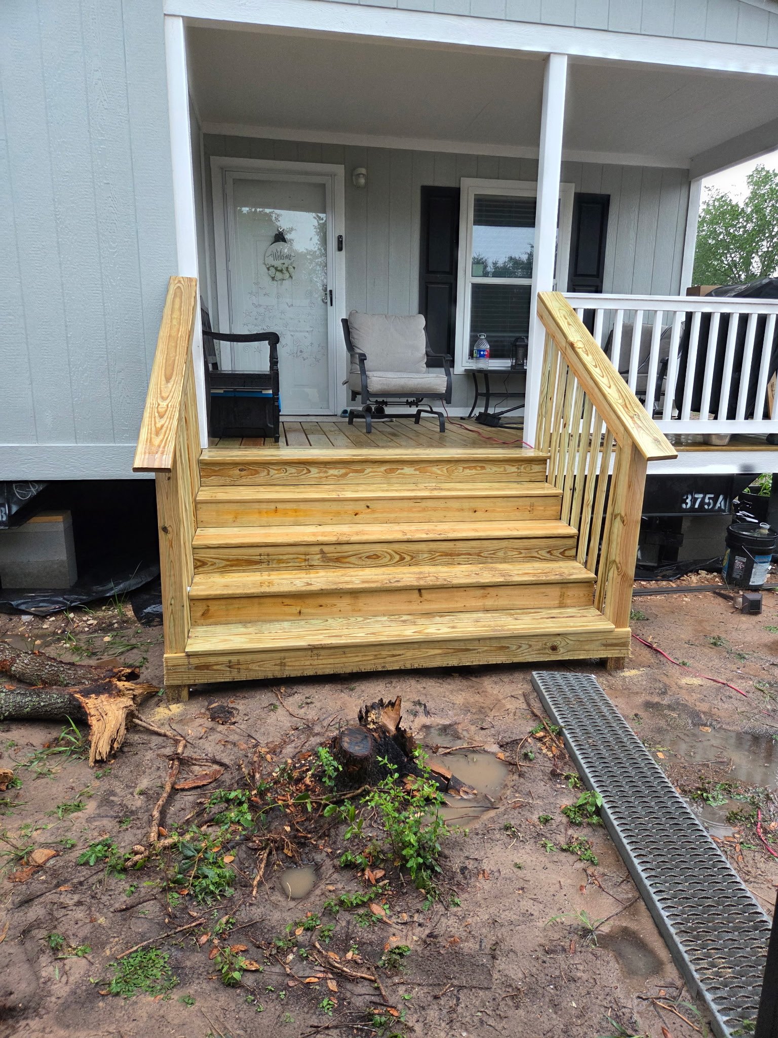 Image of a newly built wooden porch with stairs leading to a house entrance. The porch has a cushioned chair, a small side table with a water bottle, and some items on it. The surrounding ground is muddy with some scattered broken branches and green plants. Part of a drainage grate is visible in the foreground.