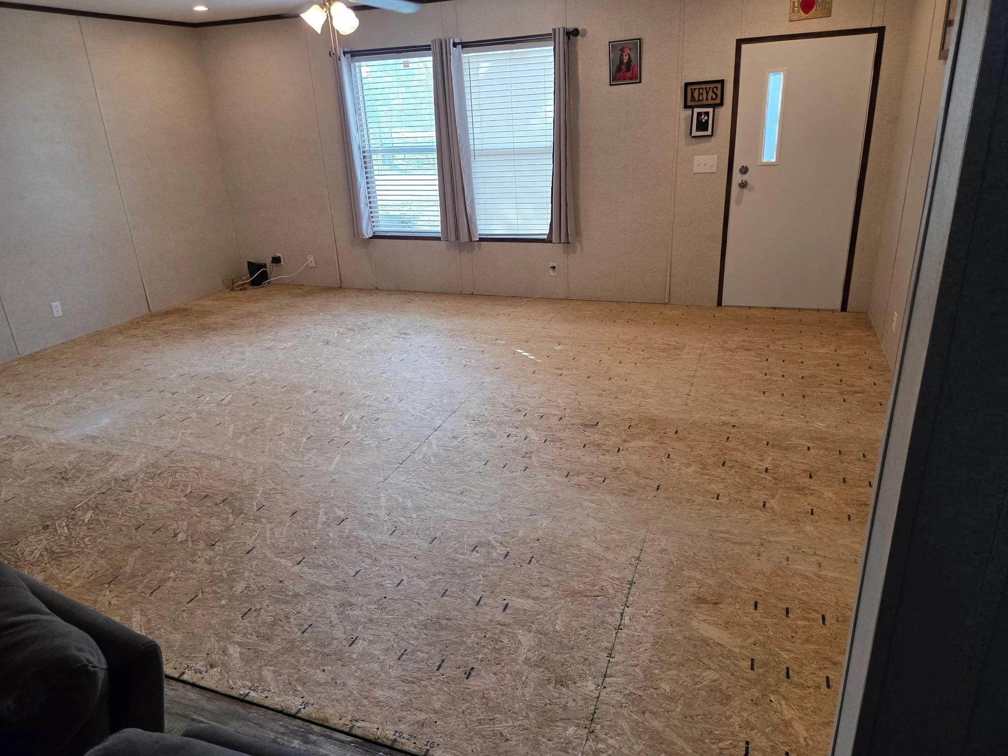 A room with a wood floor, two windows with white curtains, a ceiling fan with lights, and some wall decorations including a photo of a graduate, a sign reading 'KEYS,' and a small framed photo. There is a black leather couch partially visible in the bottom left corner.