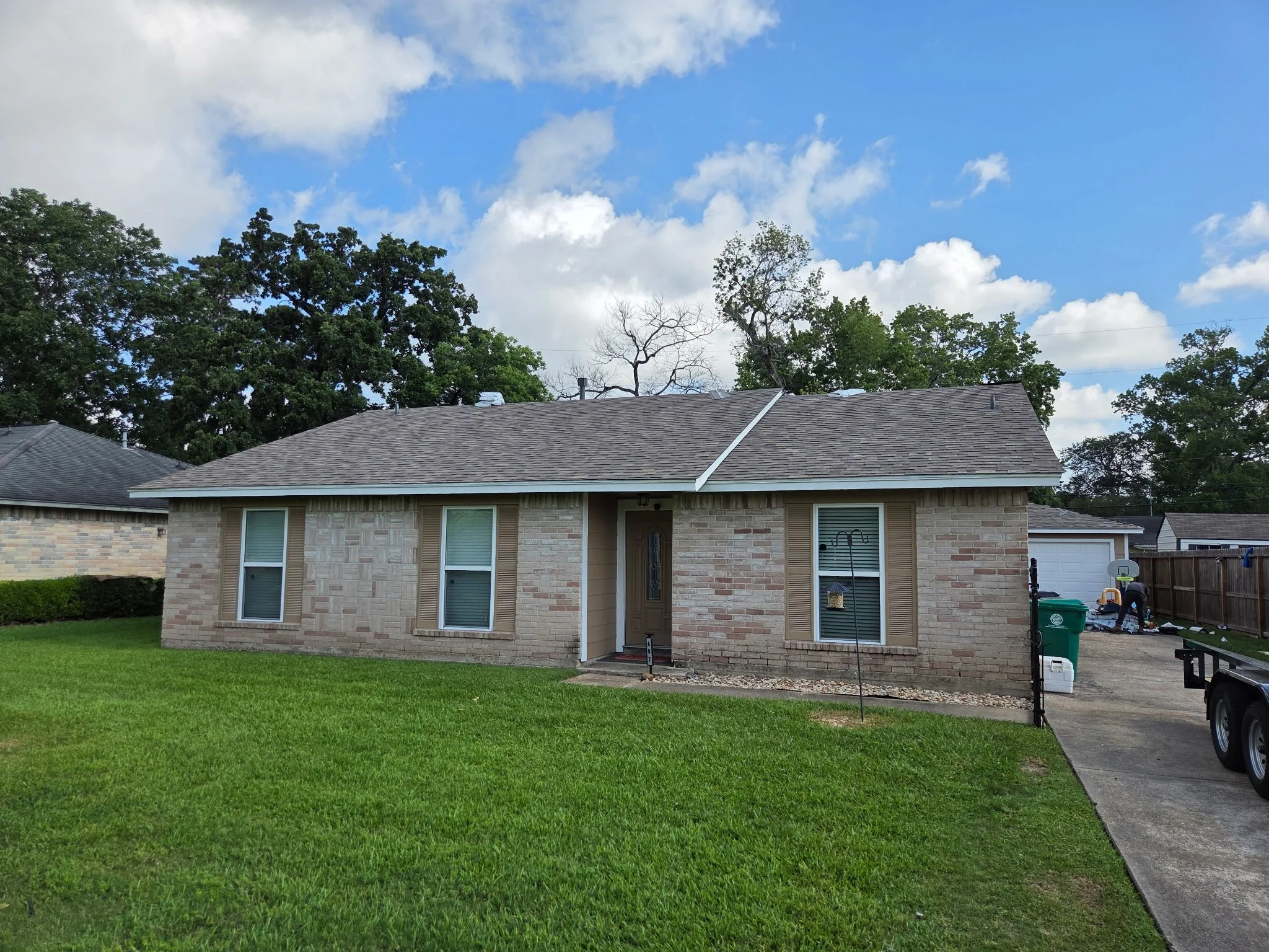 A suburban house with a brick exterior, multiple windows, a brown shingled roof, and a lawn in front. The sky above is partly cloudy with blue skies.