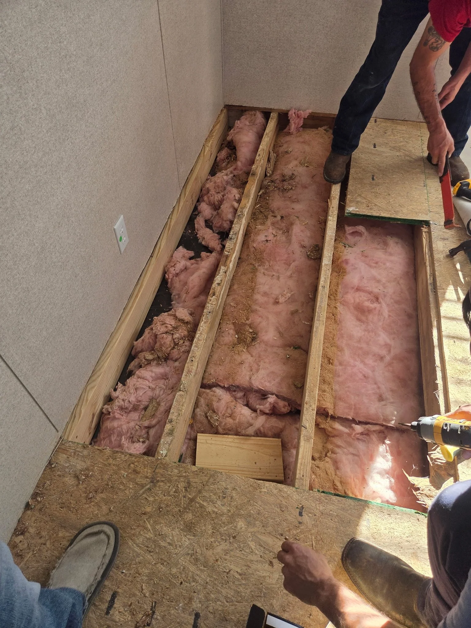 A person working on installing or repairing flooring, with insulation visible between wooden floor joists, in a room with unfinished walls and flooring.