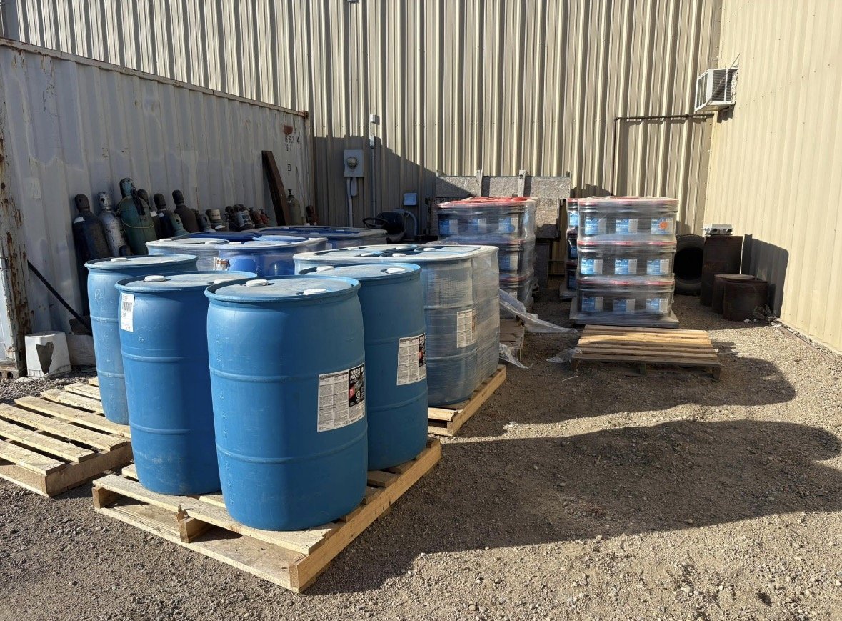 Outdoor storage area with blue barrels, pallets, and containers of chemicals against a metal fence and building wall.