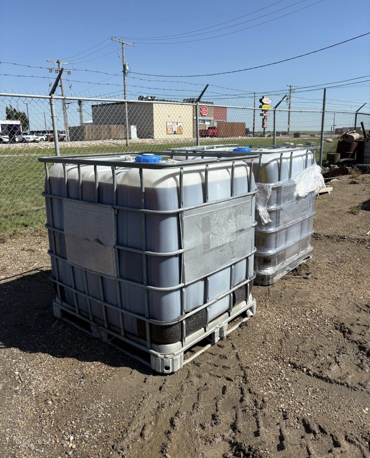 Two large white plastic water tanks on pallets, wrapped partly in plastic, outdoors on a dirt lot with a chain-link fence, power lines, and buildings in the background.