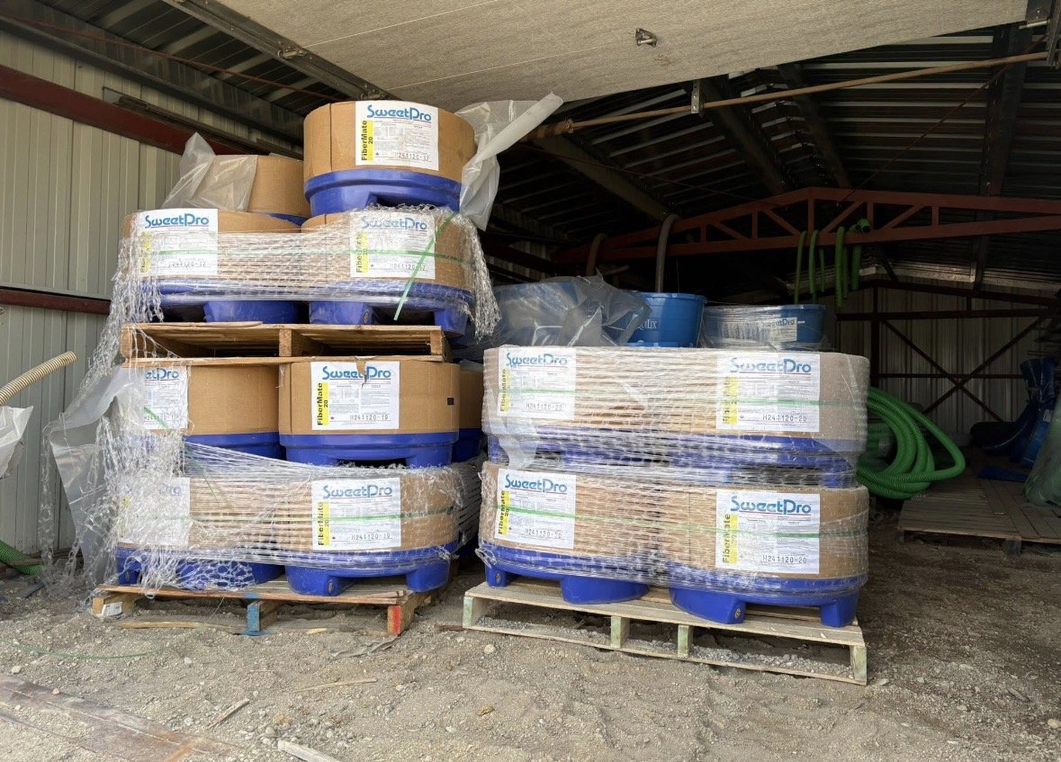 Pallets stacked with large rolls of fiber matting wrapped in clear plastic inside a storage shed.