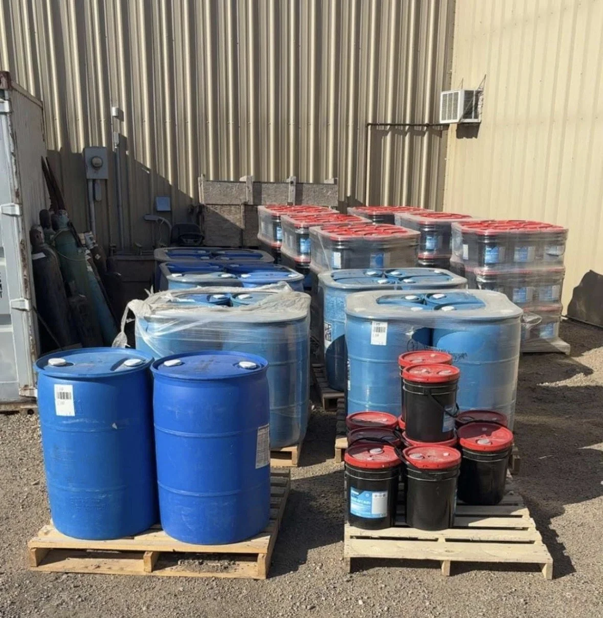Stacks of large blue and black plastic drums with red lids on wooden pallets, outside against a metal building wall.