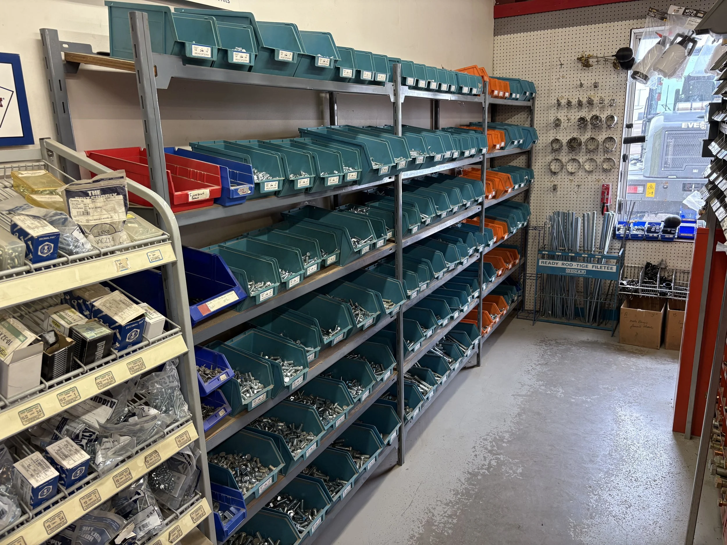 Hardware store aisle with shelves of plastic bins containing screws, nuts, and bolts. Pegboard wall with hooks holding various tools and hardware accessories. A cart with hardware supplies and a window showing a truck outside.