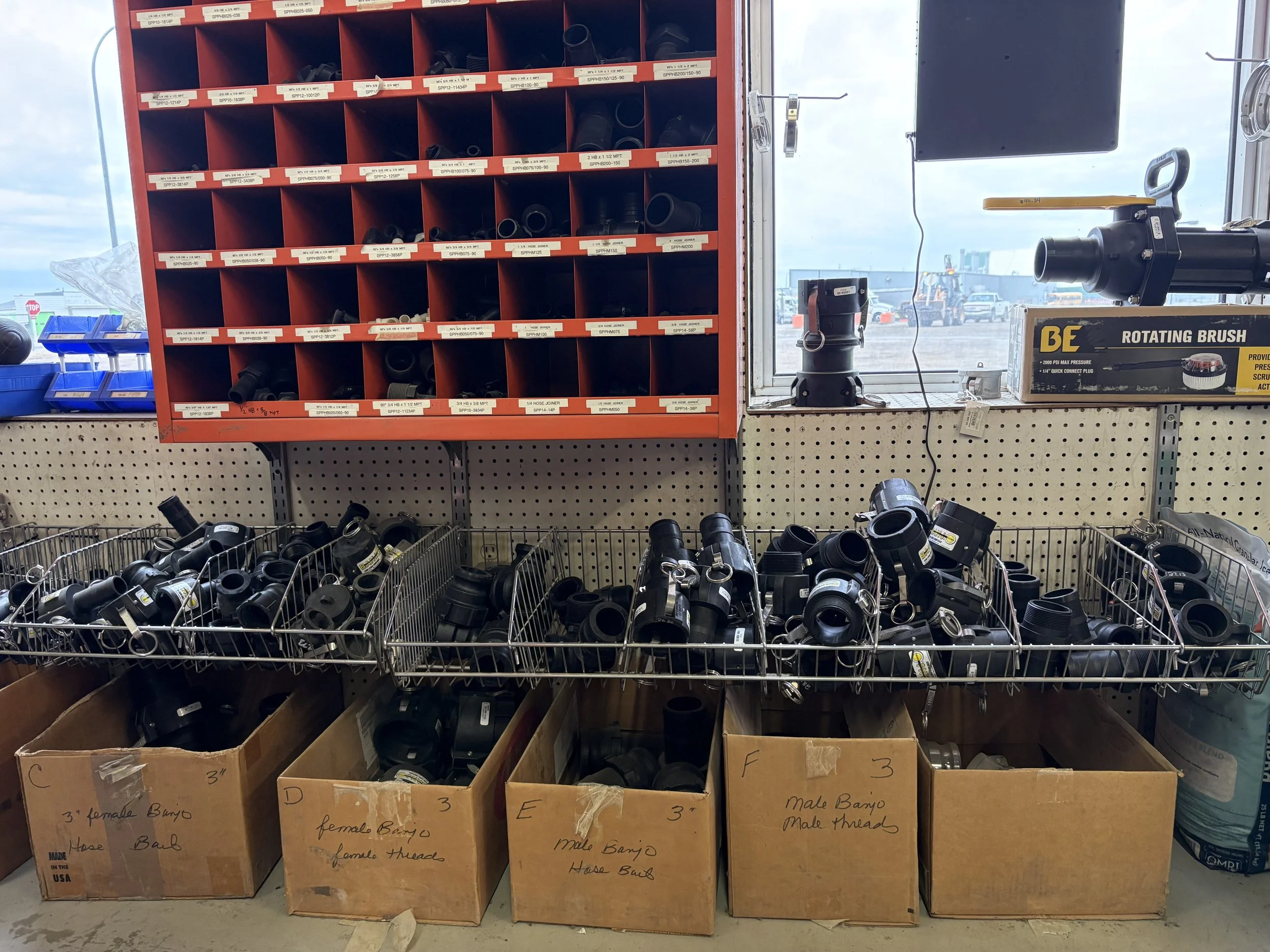 A display of various black plastic fittings and connectors arranged on red cubby shelves and in cardboard boxes on a pegboard behind a metal wire basket, inside a hardware store or warehouse.