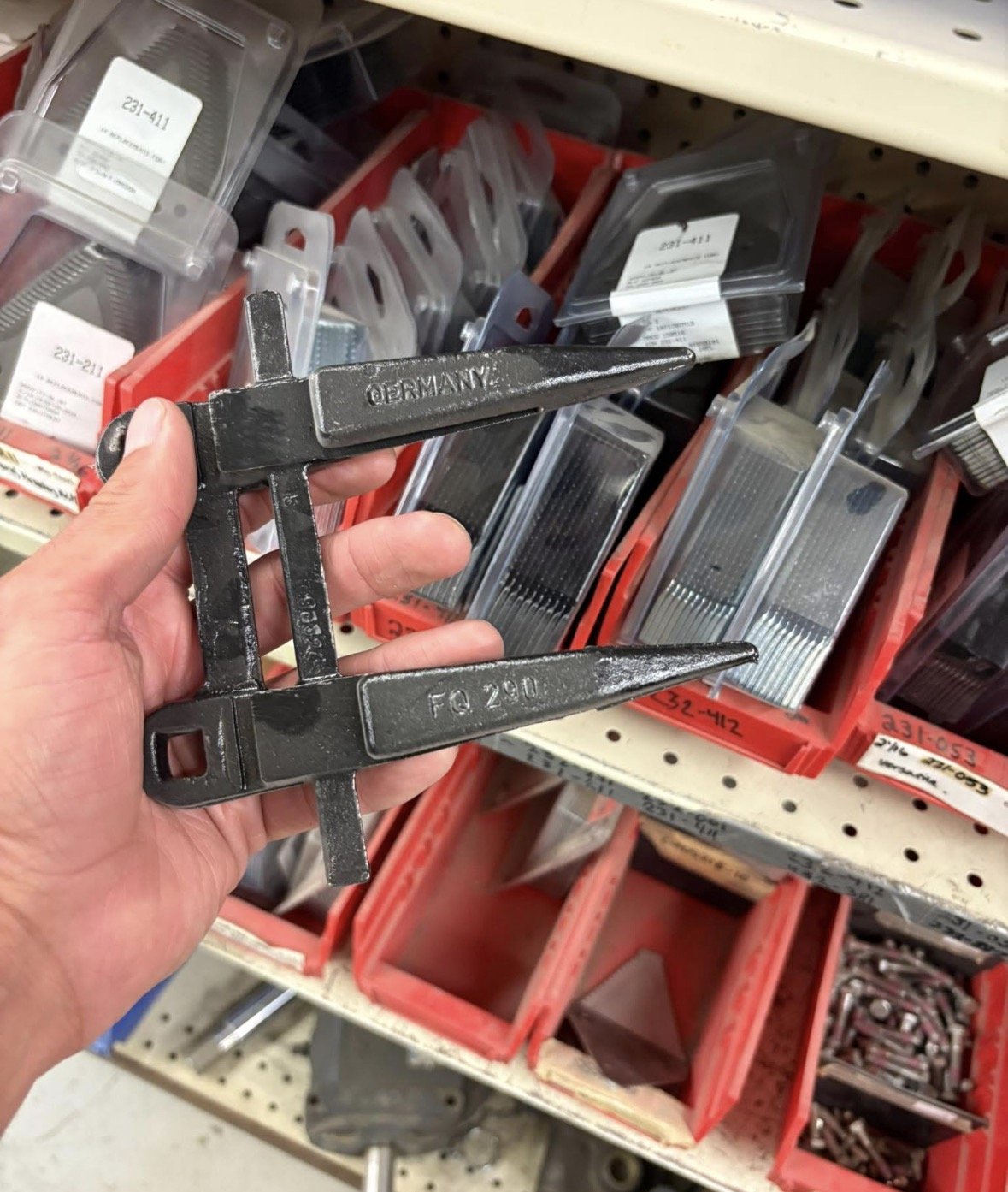 A hand holding a black, metal file clamp with the word 'GERMANY' and 'FQ 20' embossed on it. The background shows store shelves with various metal hand tools and hardware items in plastic and cardboard packaging.