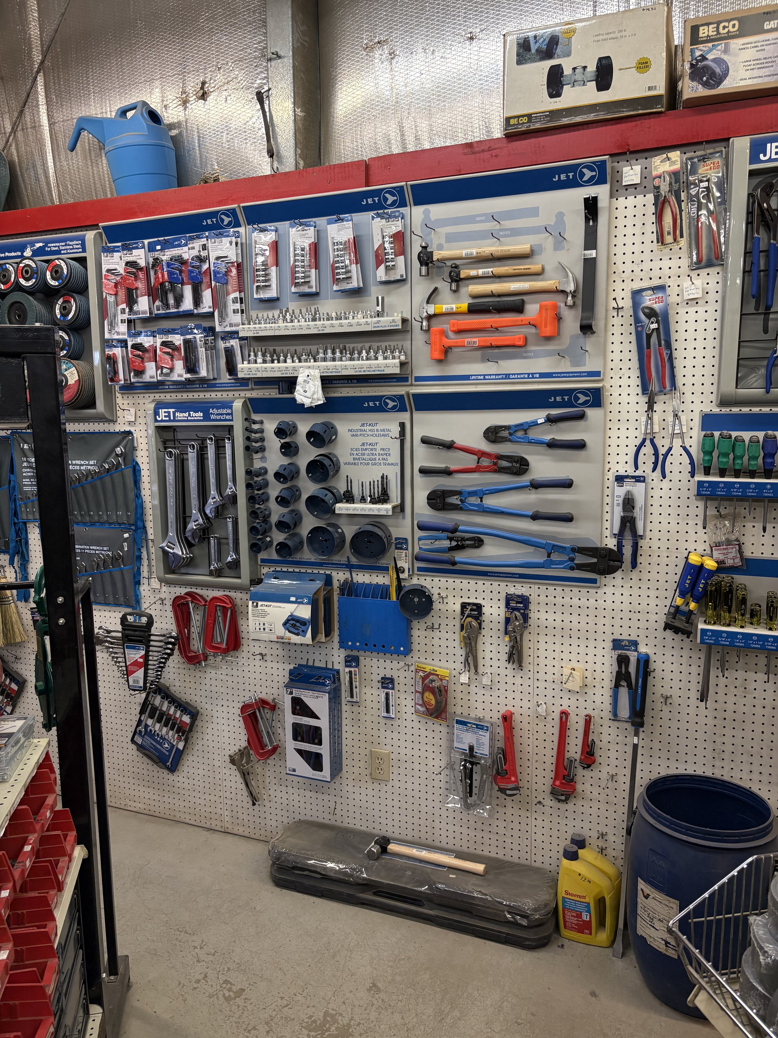Tools and hardware items arranged on a pegboard wall in a store, including wrenches, pliers, screwdrivers, hammers, and other hand tools, with some boxes and containers on the floor and shelves.