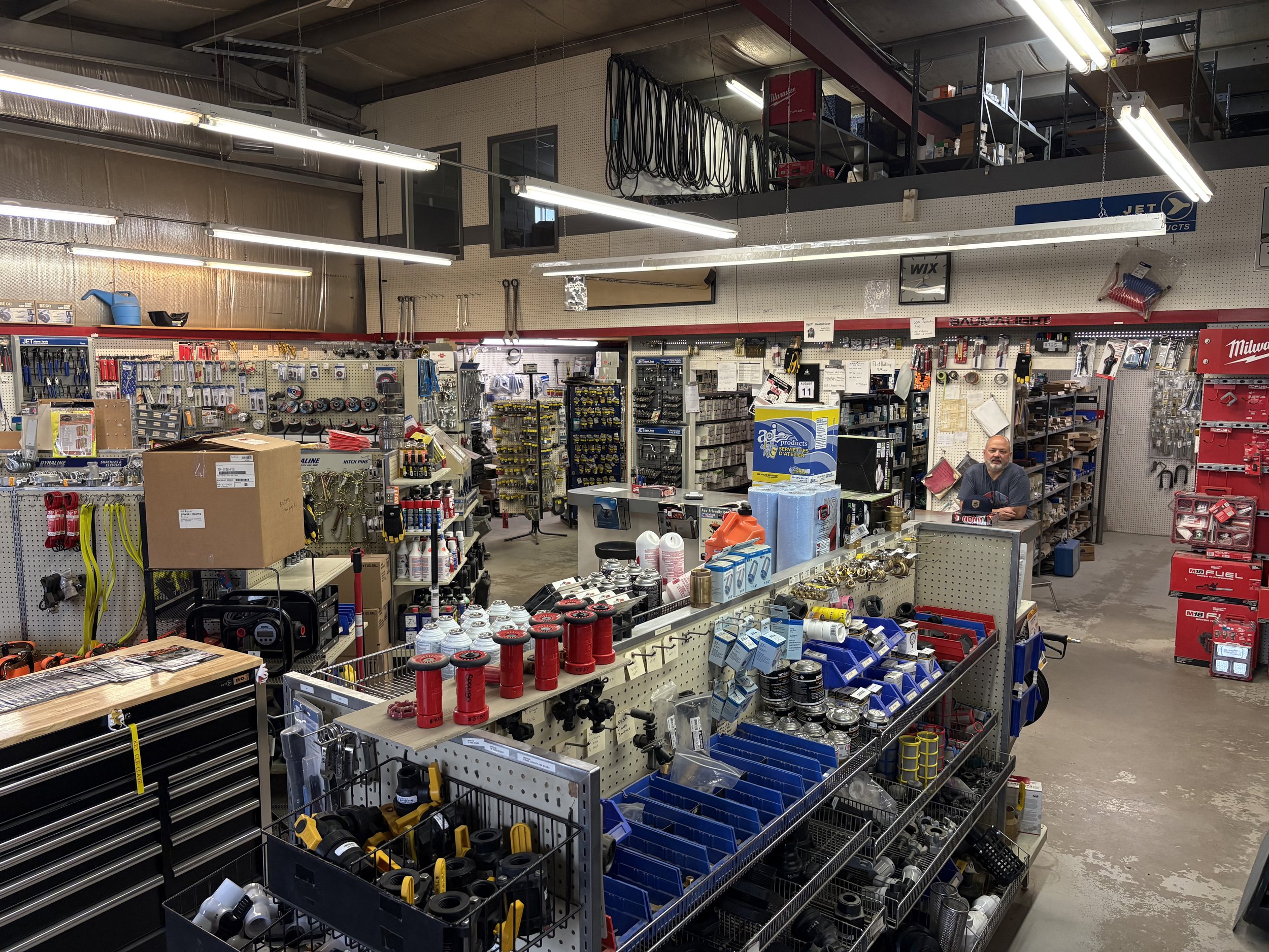 Interior of a hardware store with shelves stocked with tools, hardware supplies, and accessories. A man is standing behind the counter in the lower right corner.