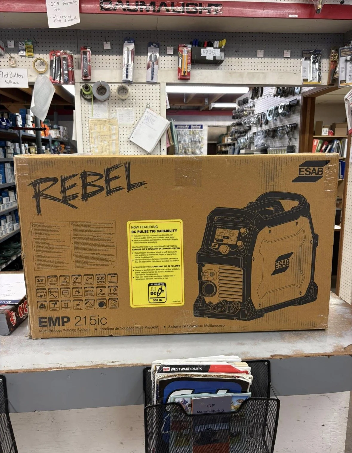 A boxed ESAB Rebel EMP 215IC multi-process welding system placed on a display counter in a hardware store. Behind the box, there are tools, tapes, and signage on a pegboard wall.