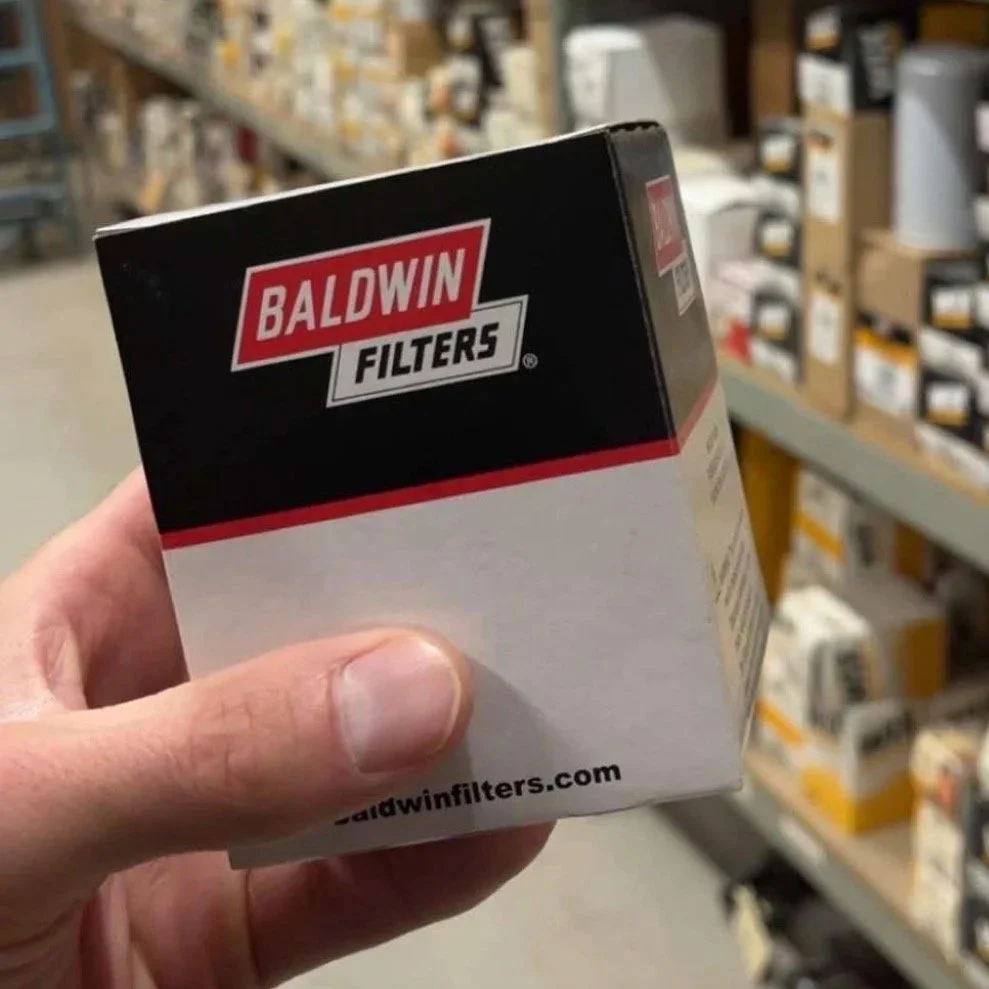 Hand holding a Baldwin Filters box in auto parts store aisle with shelves of other auto parts and filters in the background.