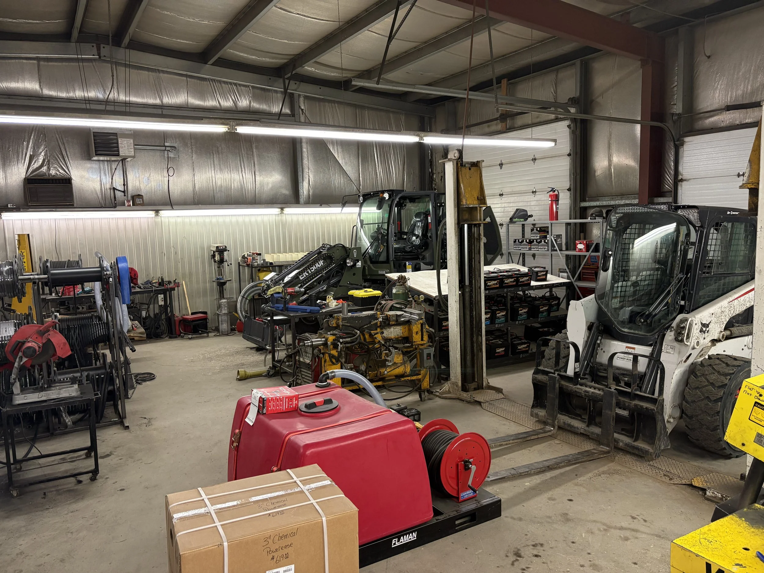 Interior of a workshop or garage with various tools, machinery, and equipment, including a small construction vehicle, shelves with batteries, a fire extinguisher, and cardboard boxes, with metallic walls and ceiling.