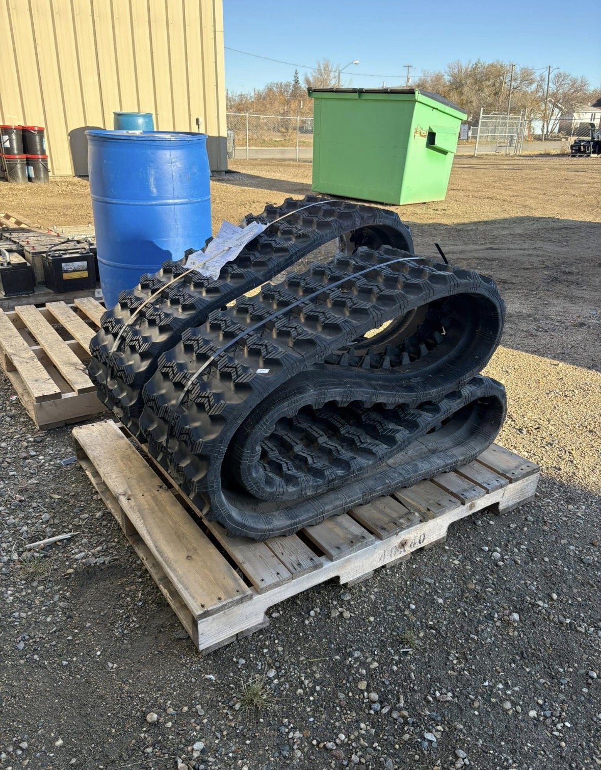 Two black rubber tracks with a tread pattern, stacked on a wooden pallet outdoors. A blue plastic barrel and a green storage container are visible in the background.