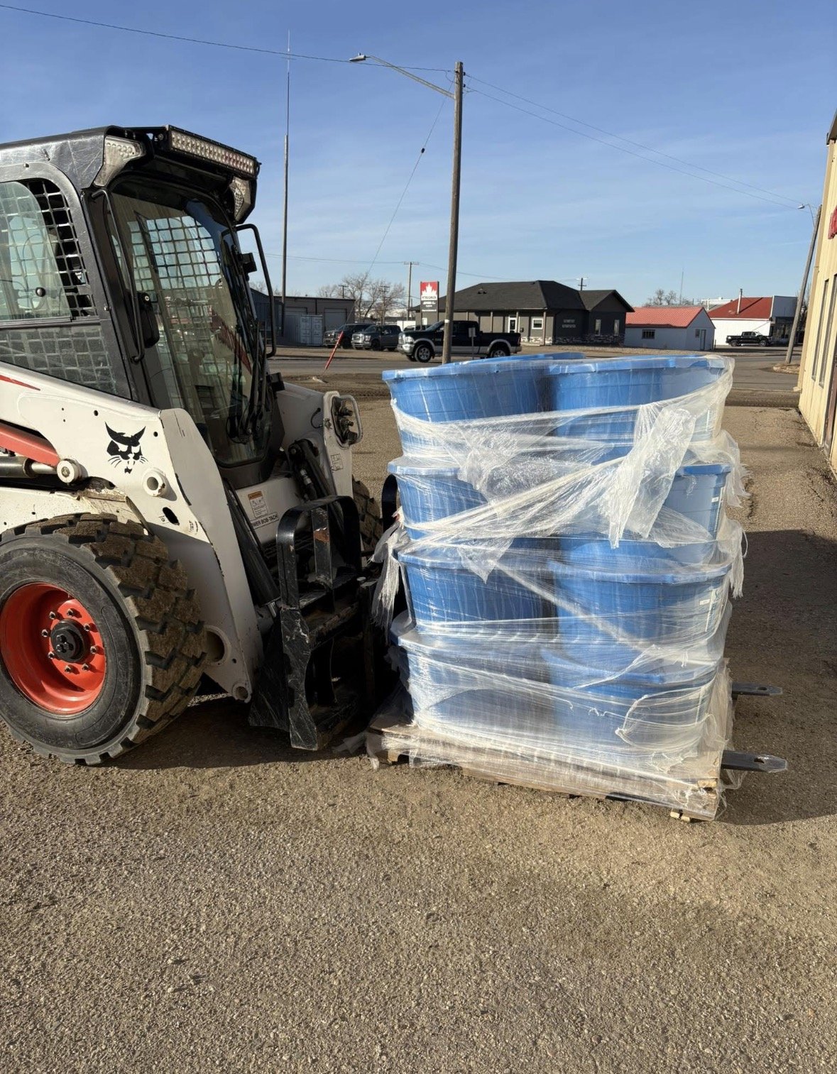 A skid steer loader with a black and white body, red wheels, and a lion logo on the side, holding a pallet of large blue plastic tubs wrapped in clear plastic film, outdoors on a dirt lot, with buildings and utility poles in the background.