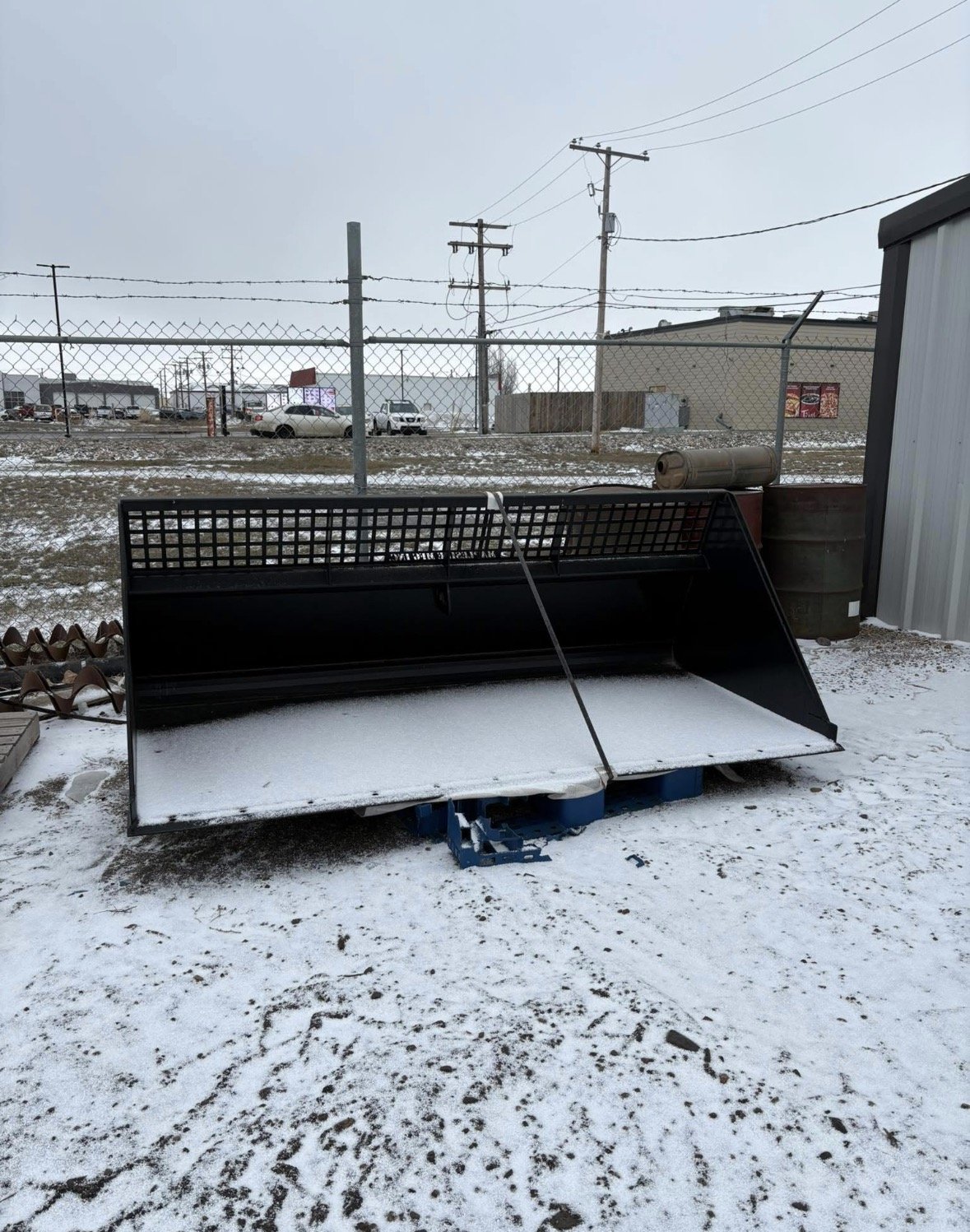 Snow-covered construction equipment and tools outdoors, surrounded by a chain-link fence and industrial buildings in the background.