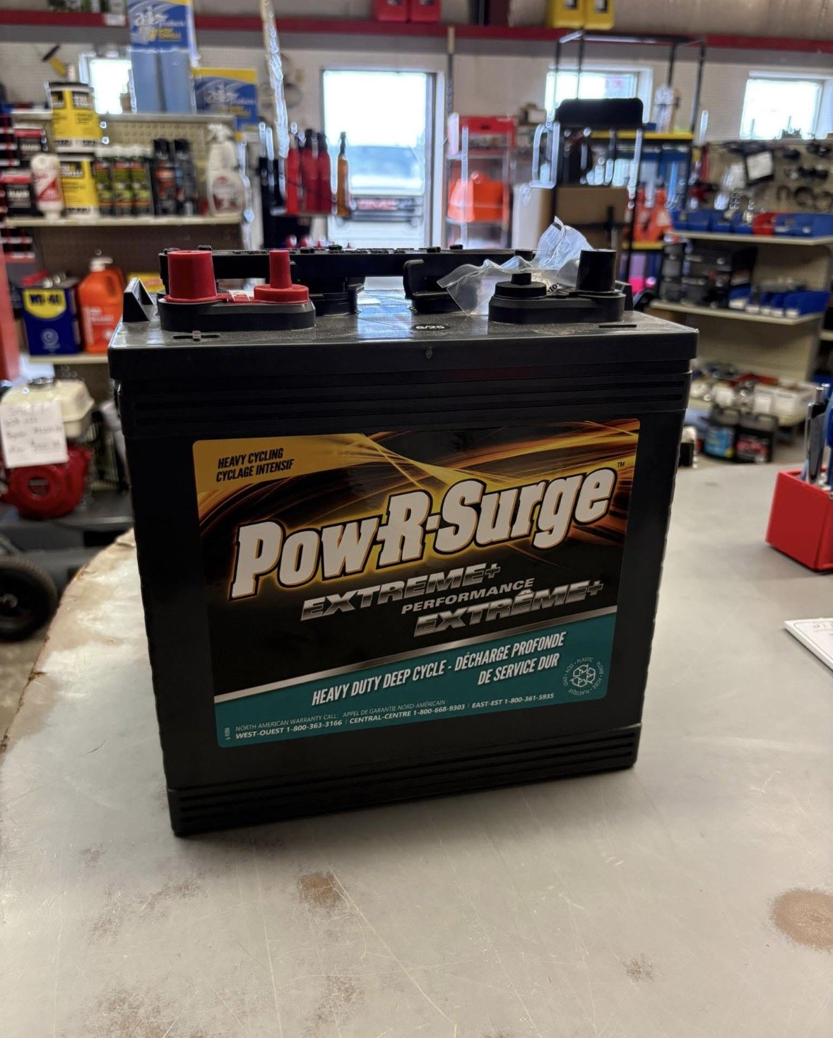 A black car battery labeled "Pow R Surge Extreme+" on a workbench in a hardware store, with shelves of tools and supplies in the background.