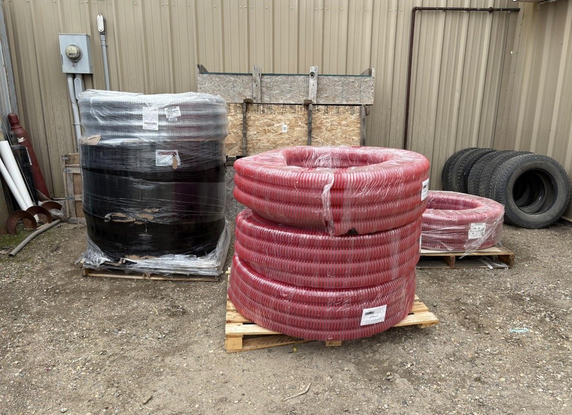 Stacks of red and black coiled hoses, some wrapped in plastic, placed on wooden pallets outside on dirt ground. There is a beige metal wall in the background and tires to the right.