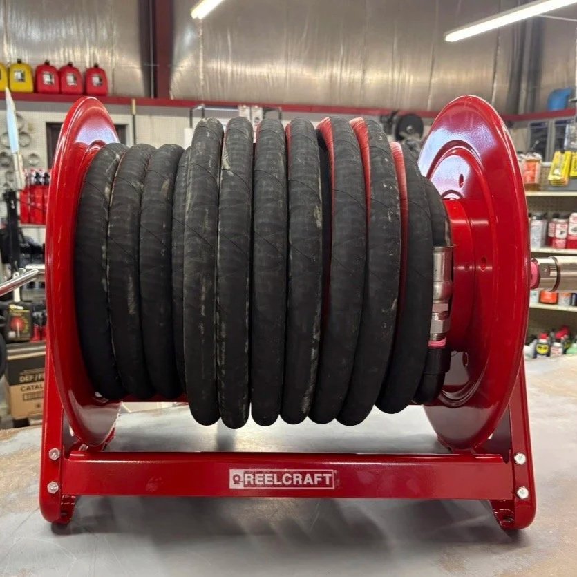 A red industrial reel cart holding multiple black rubber hoses stacked around a metal spool, with various tools and supplies in the background.