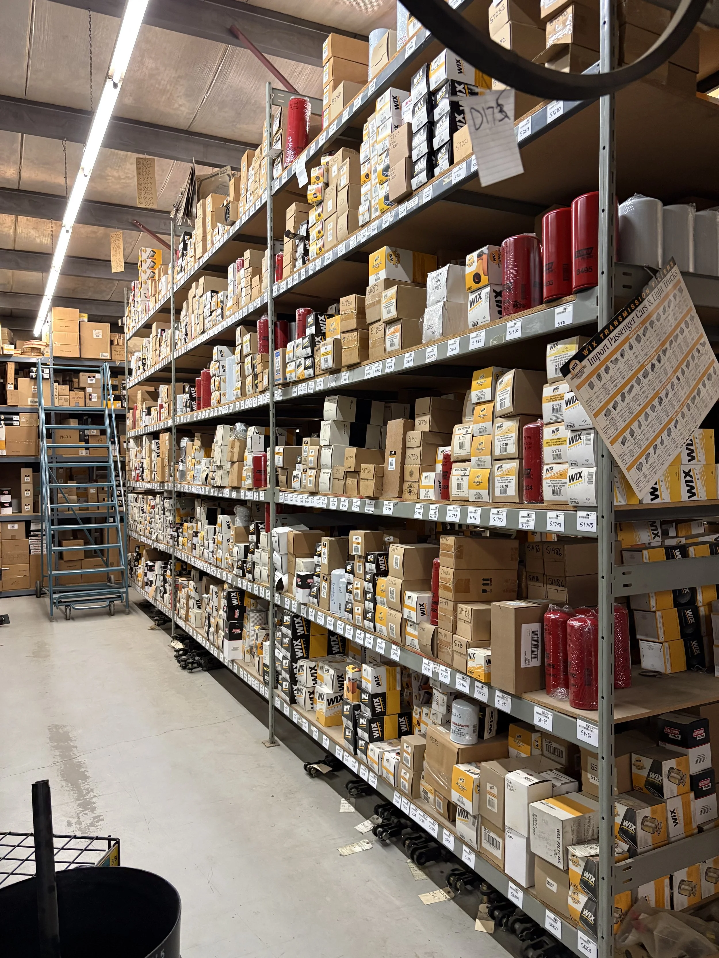 A section of a warehouse aisle with metal shelves filled with boxes of automotive parts, including filters. A blue rolling ladder is positioned along the aisle. The ceiling features fluorescent lighting and exposed beams.