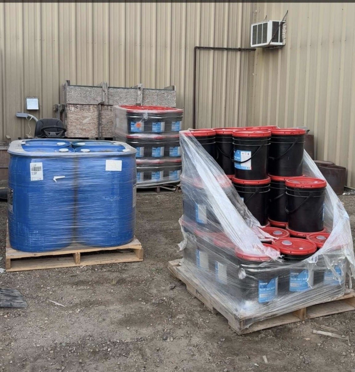 Outdoor storage of large water tanks and buckets wrapped in plastic pallets against a beige metal building wall.