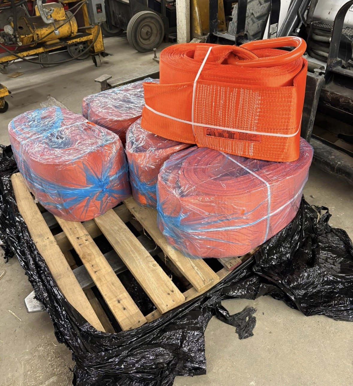 Orange safety traffic cones and orange safety straps on a wooden pallet, wrapped in clear plastic, in an industrial or warehouse setting.