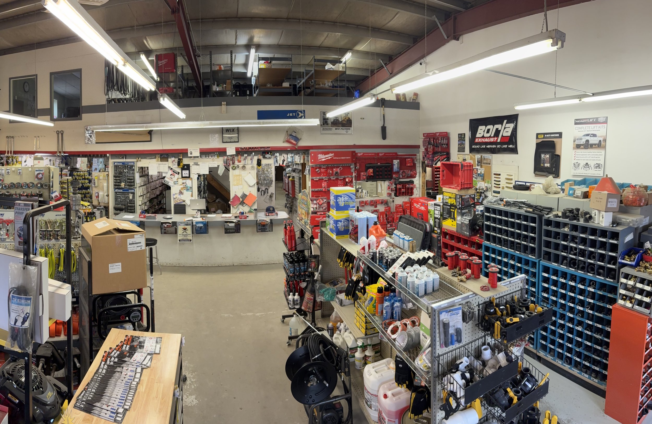 Interior of a hardware store with shelves stocked with tools, supplies, and equipment, including spray cans, power tools, and storage bins, with a counter and cardboard boxes in the foreground.