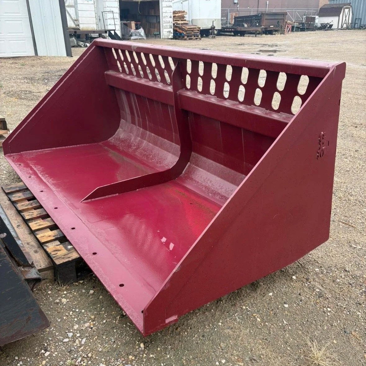 Red agricultural loader bucket with a reinforced cutting edge, situated outdoors on a gravel surface, with industrial buildings and equipment in the background.